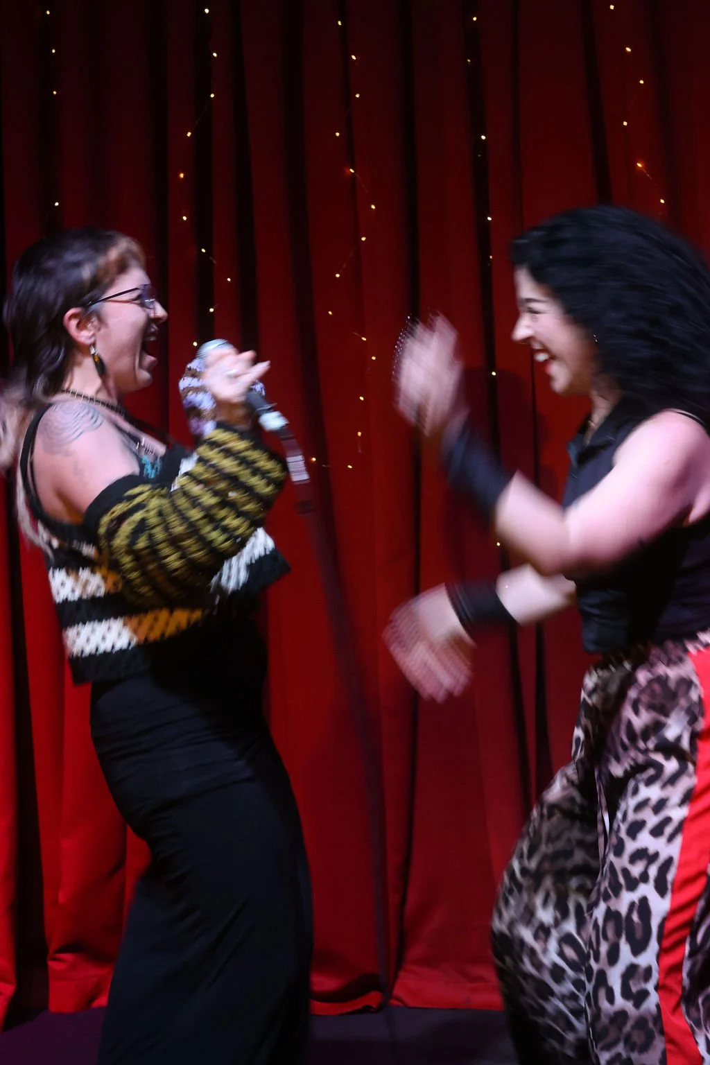 Two women with dark hair laughing and celebrating on stage in front of a red curtain with string lights. One is holding a microphone, and both are wearing colorful, patterned outfits.