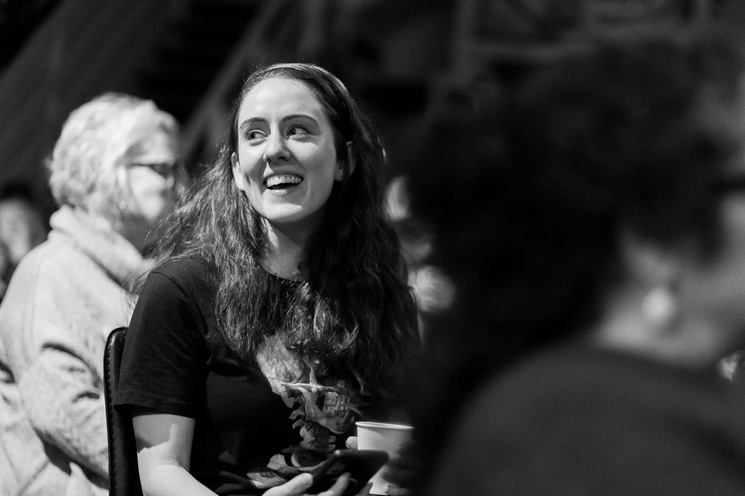 A young woman with long wavy hair smiling and looking to her left, holding a cup in her left hand, sitting in a crowded place with other people in the background.