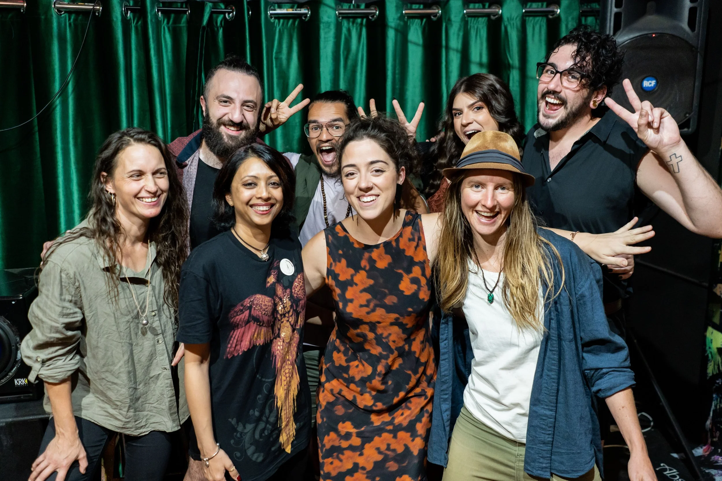A group of ten diverse people are smiling and posing together in front of a green curtain, some making peace signs, at what appears to be a celebration or gathering.