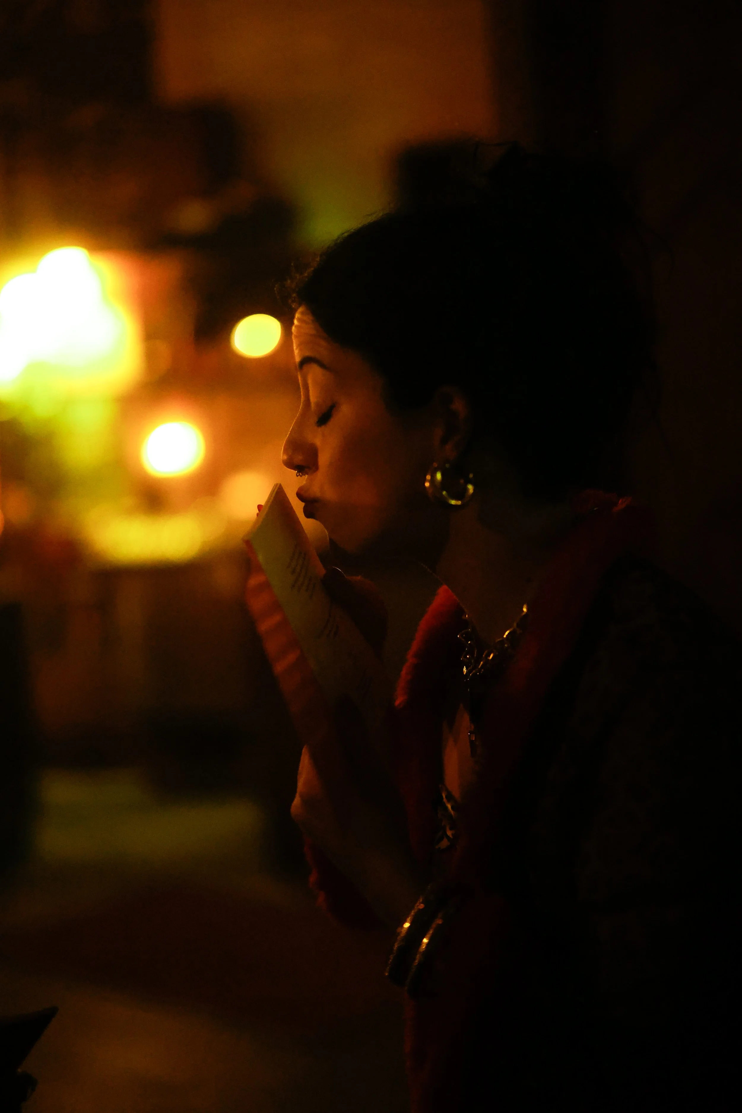 A woman with a bun hairstyle, gold hoop earrings, and dark clothing is reading a piece of paper in a dark, nighttime setting illuminated by warm, out-of-focus lights.