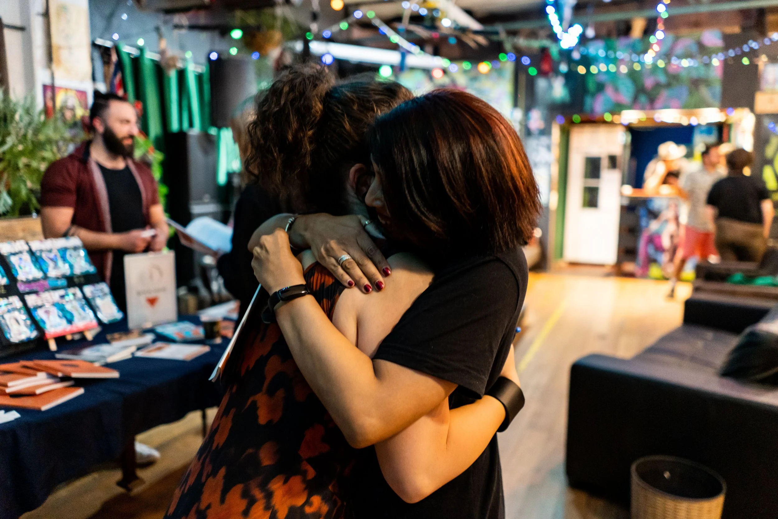 Two women hugging each other at a lively indoor event with colorful string lights and a marketplace or expo setting in the background.