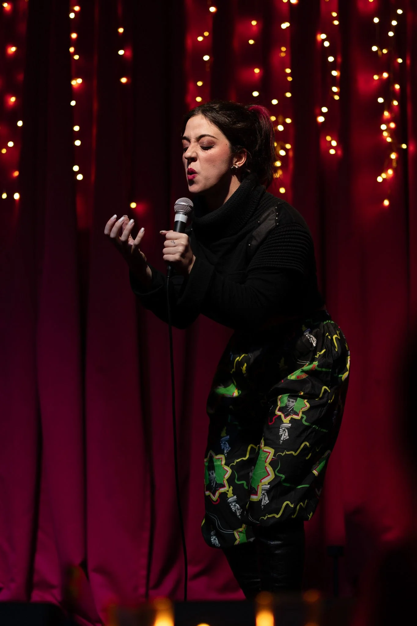 Woman performing on stage with red curtain and fairy lights in the background.