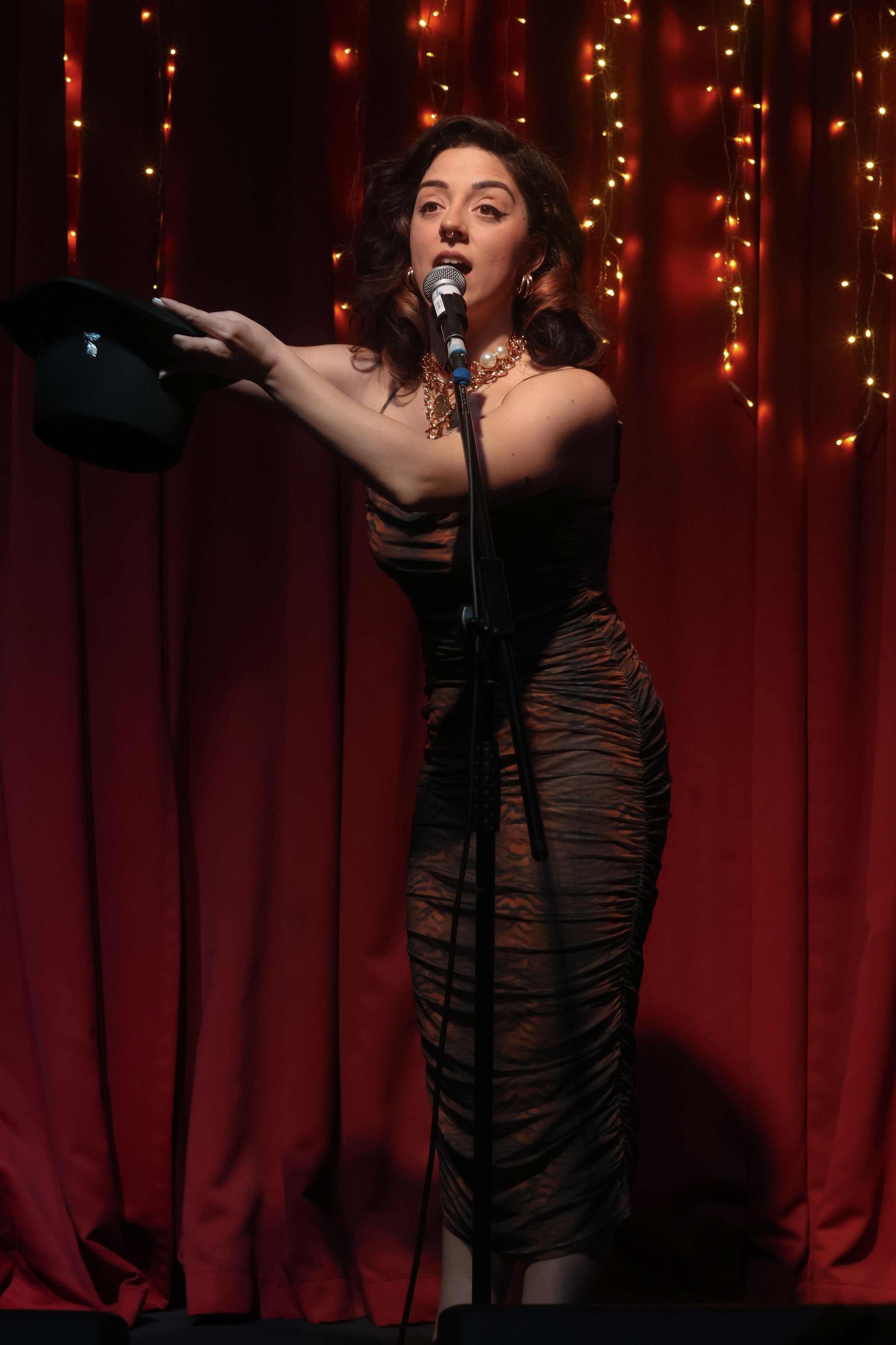 A woman with dark hair wearing a patterned dress, pearl necklace, and hoop earrings, holding a black hat in her left hand, singing into a microphone on a stage with red curtains and string lights in the background.