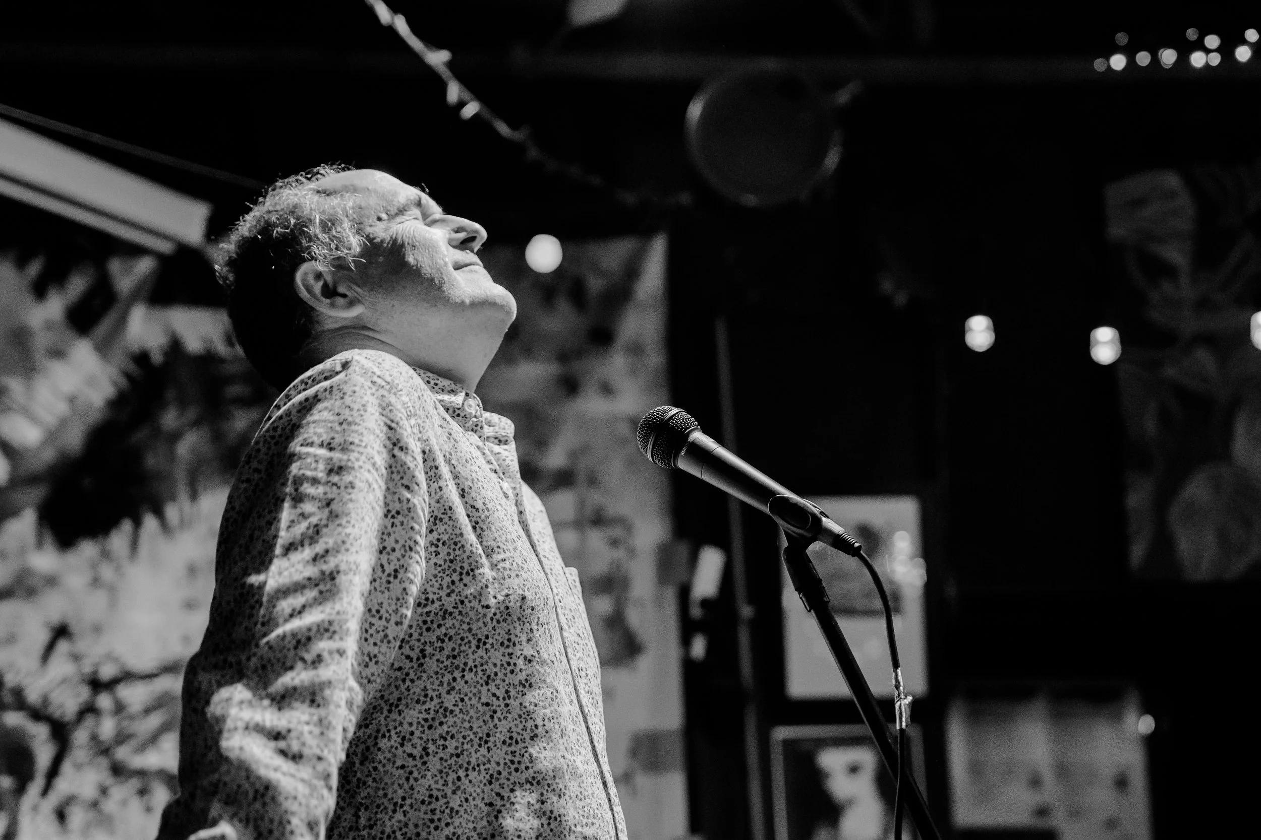 An elderly woman singing into a microphone on stage with her eyes closed and a joyful expression.