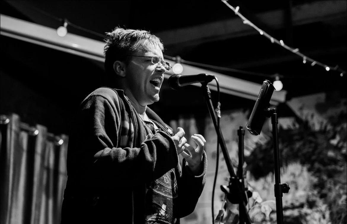 Black and white photo of a person singing passionately into a microphone, with their hands gesturing, on a stage with string lights overhead.