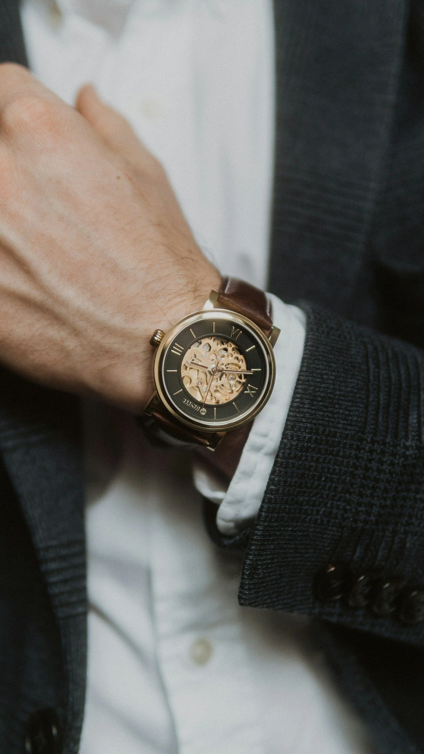 A man wearing a brown leather strap watch with a skeleton dial, dressed in a white shirt and a dark blazer.