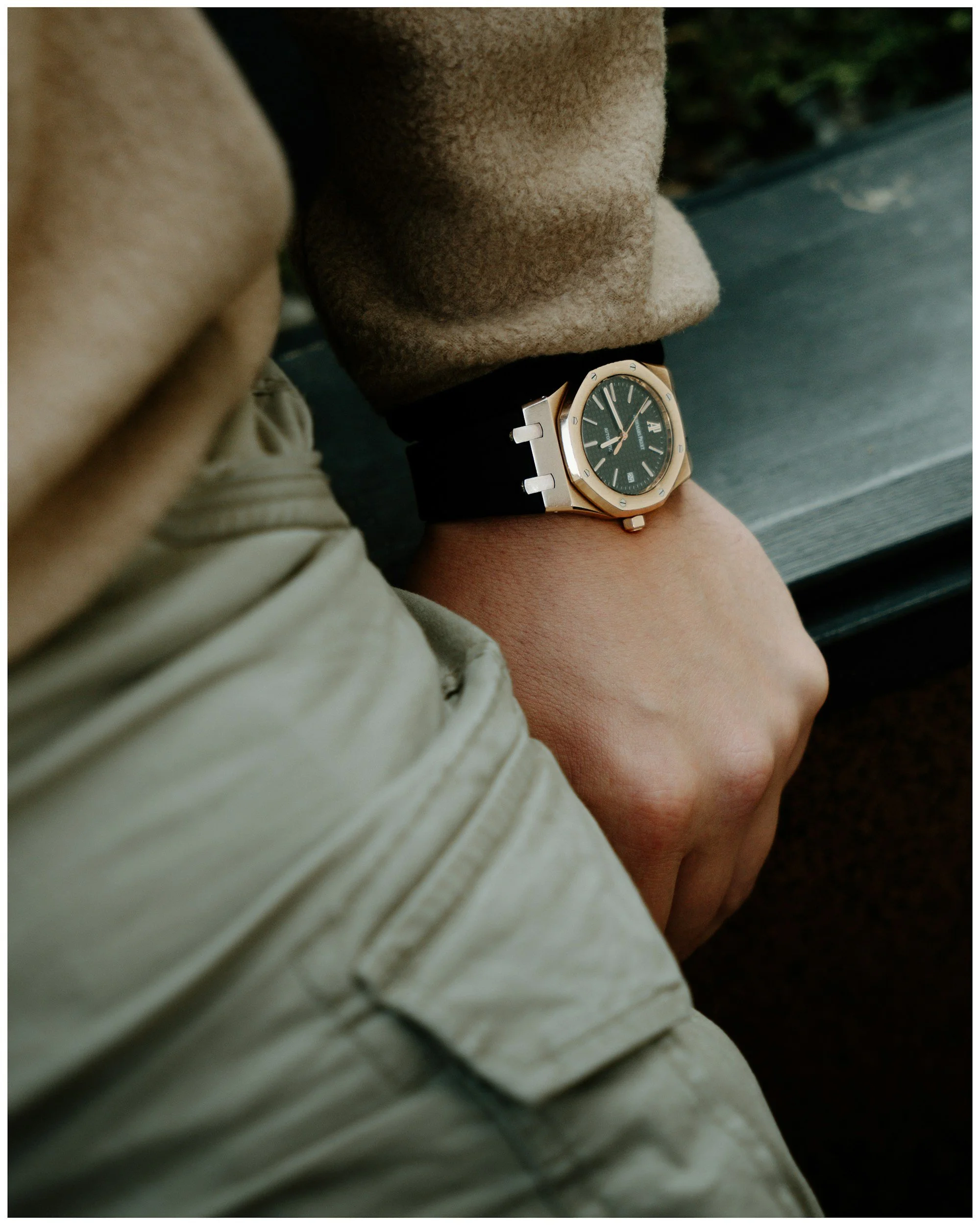 Close-up of a person's wrist with a gold and black watch, wearing a beige or brown coat, sitting outdoors on a dark surface.