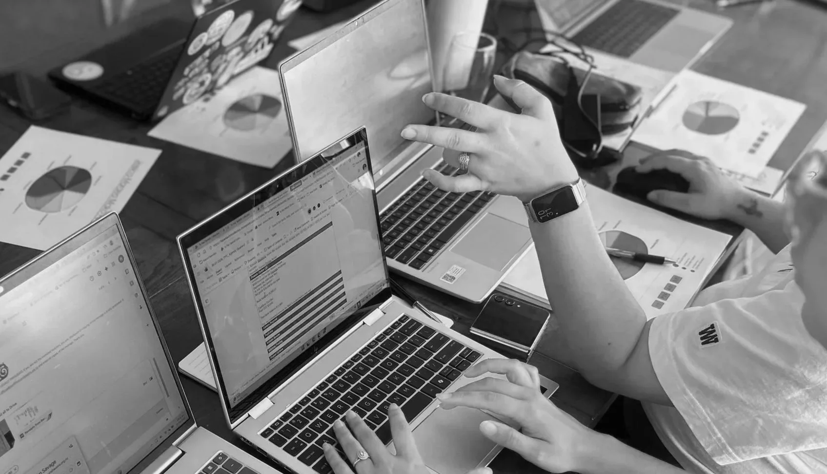 A black and white photo of a workspace with multiple laptops, documents with pie charts, a smartphone, a pen, and a smartwatch on a person's wrist. Several hands are shown interacting with the laptops.