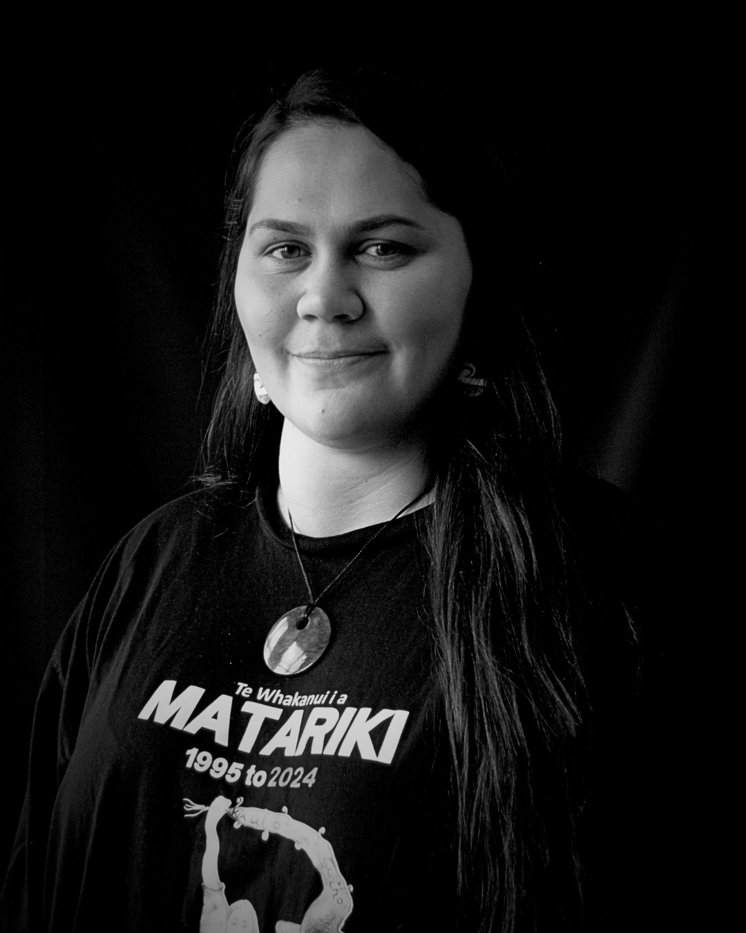 Black and white portrait of a woman wearing a T-shirt with the word 'Matariki,' indicating the Māori New Year, and a necklace with a circular pounamu pendant.
