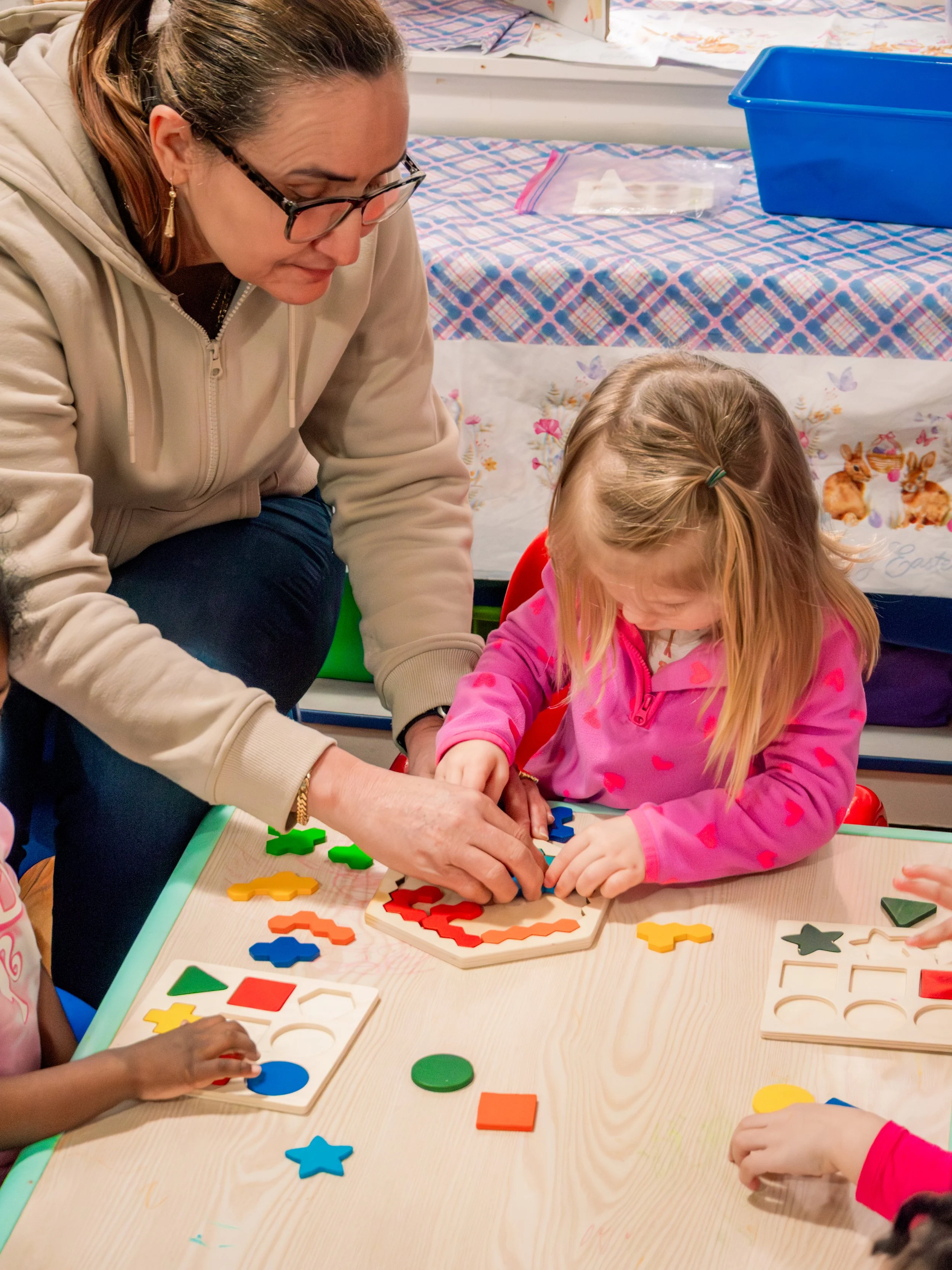 Two young girls and an adult woman playing with educational puzzle shapes at a table.