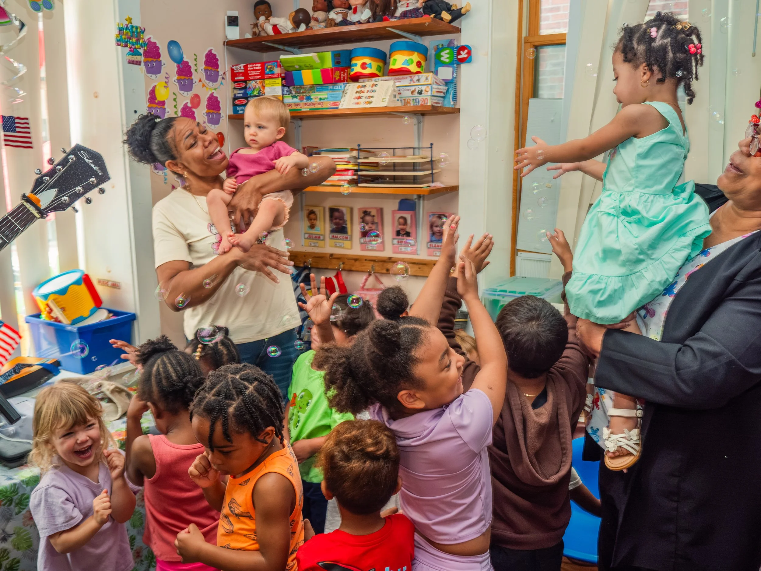 Children and adults celebrating a birthday in a decorated room with toys, balloons, and colorful decorations, including a birthday banner and cupcake wall decal.