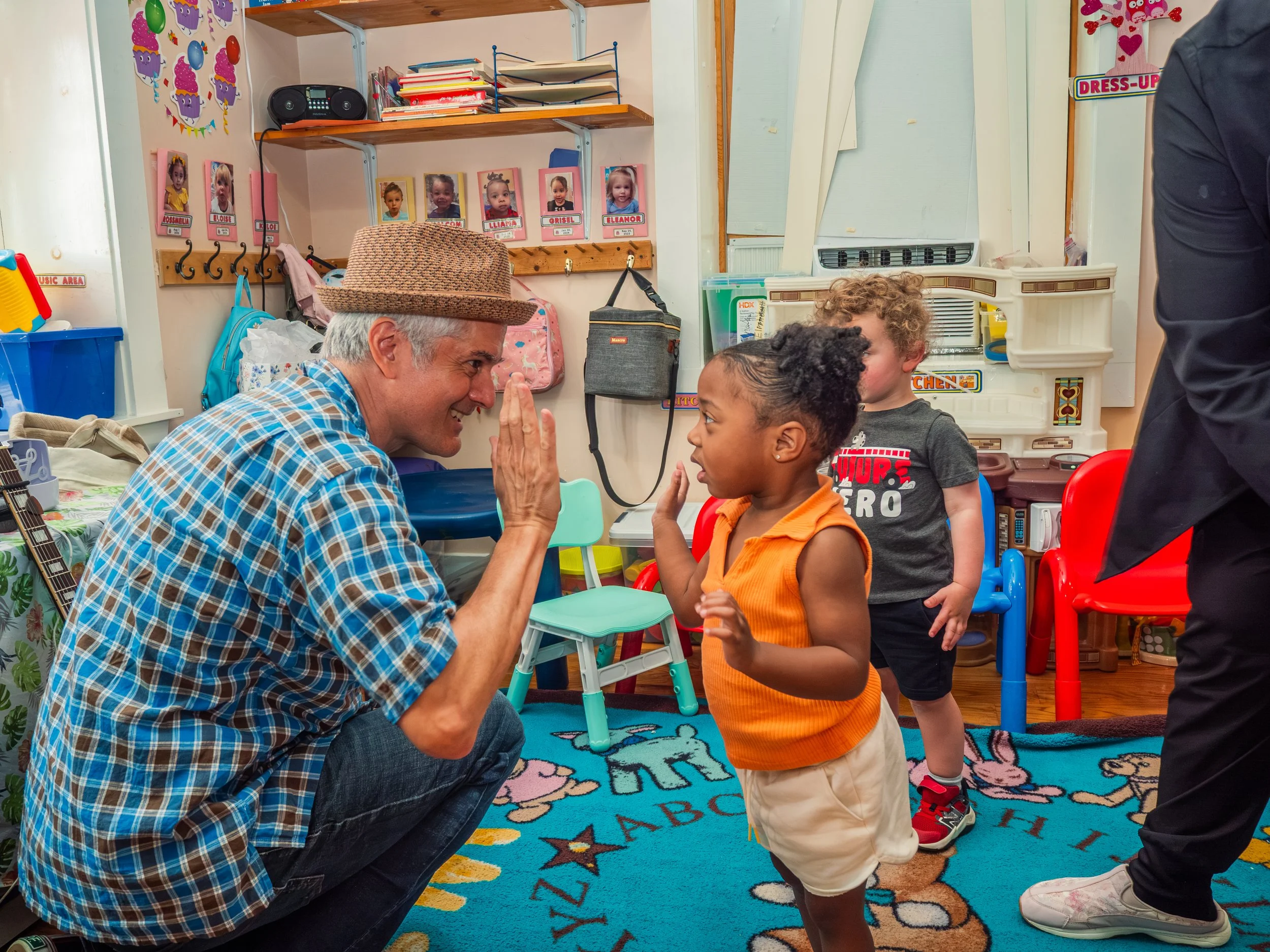 A man with a hat kneeling and smiling at a young boy in an orange shirt and khaki shorts, engaging in a high-five. Two other children are present in the background, and various colorful decorations and furniture are visible in the indoor classroom setting.