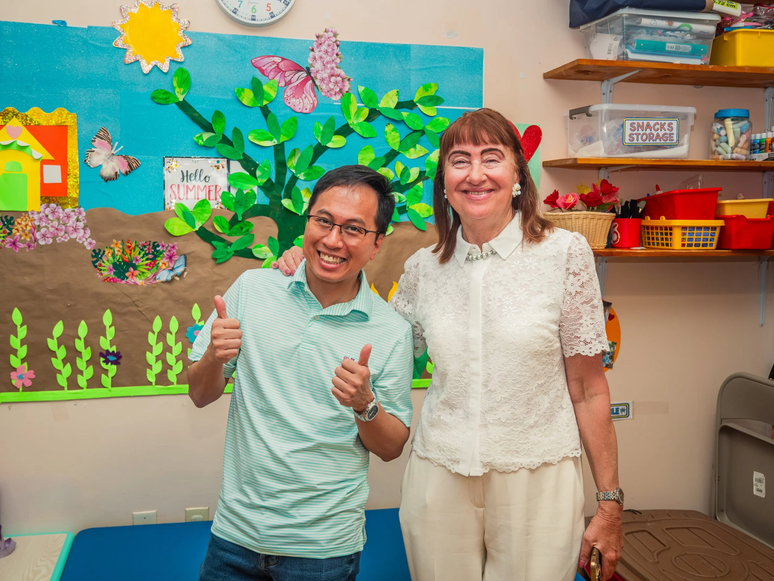 Two people smiling and giving thumbs up in front of a colorful summer-themed classroom wall display with flowers, butterflies, and a sign that reads 'Hello Summer'.