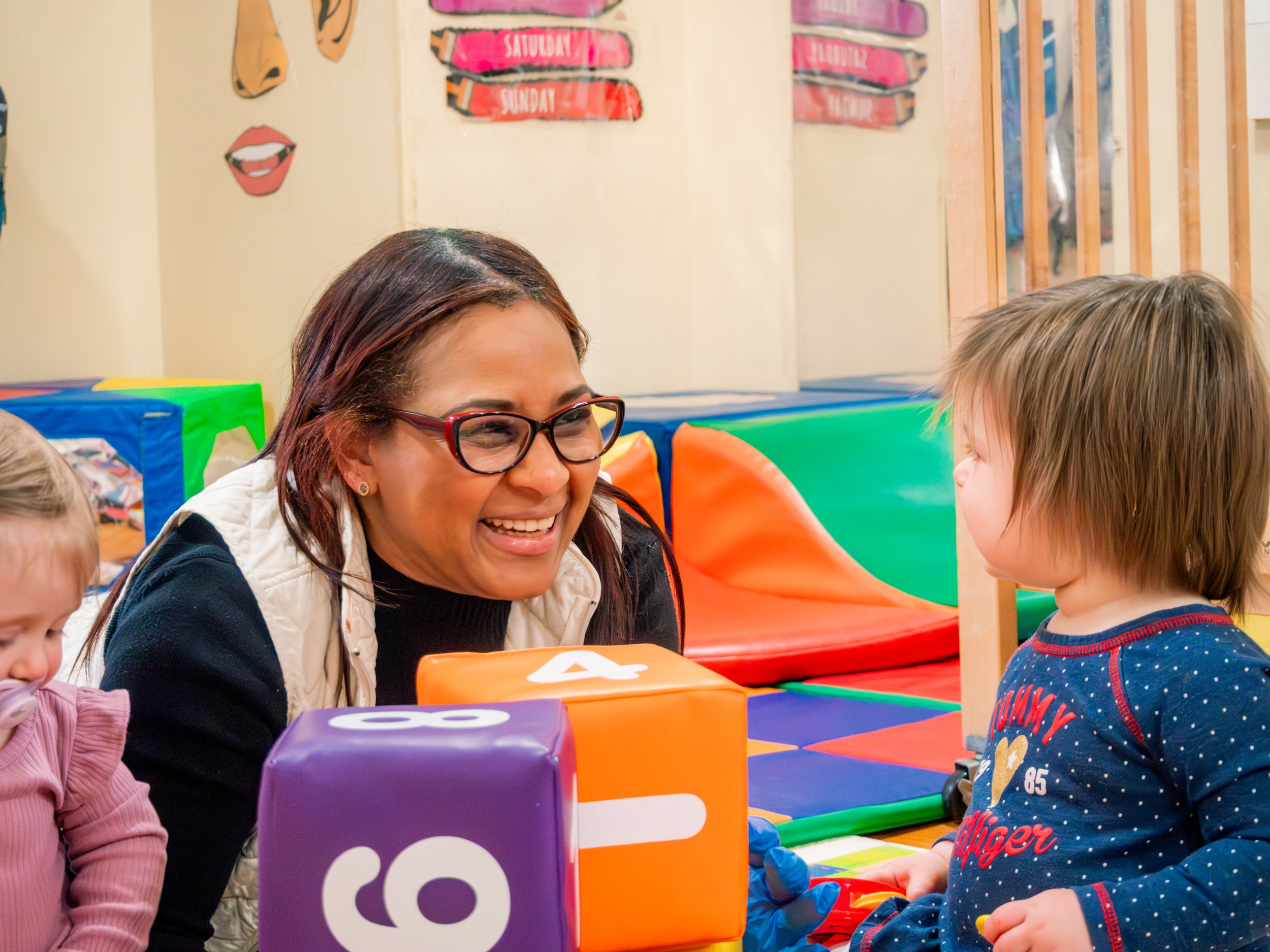 A woman with glasses smiling and talking to a young boy wearing a blue and red shirt, sitting at a table with colorful blocks and toys in a children's play area or classroom.