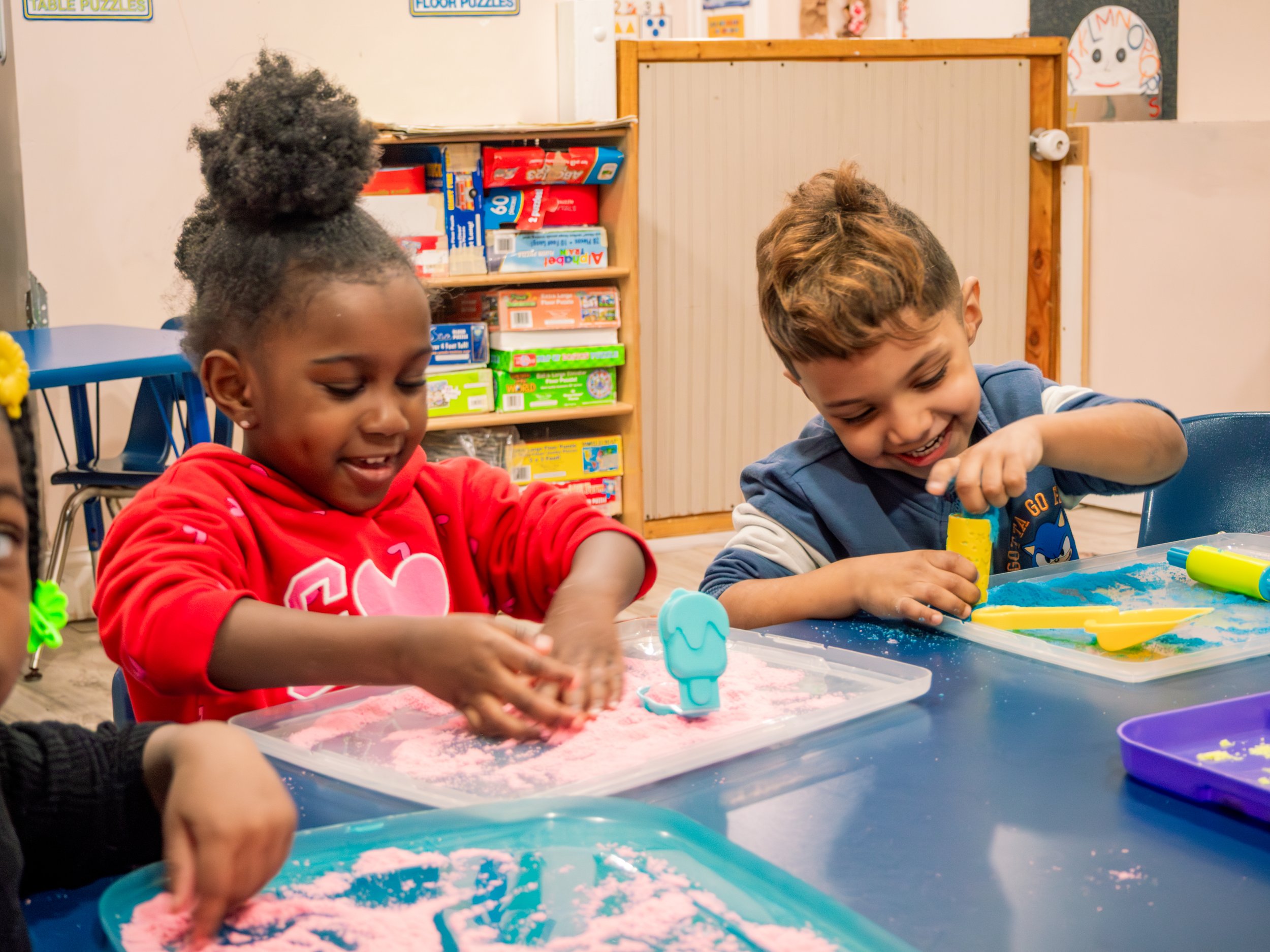 Two children, a girl with curly hair in a red hoodie and a boy with short light brown hair in a blue and white shirt, smile and play with colorful sand tools at a table in a classroom.