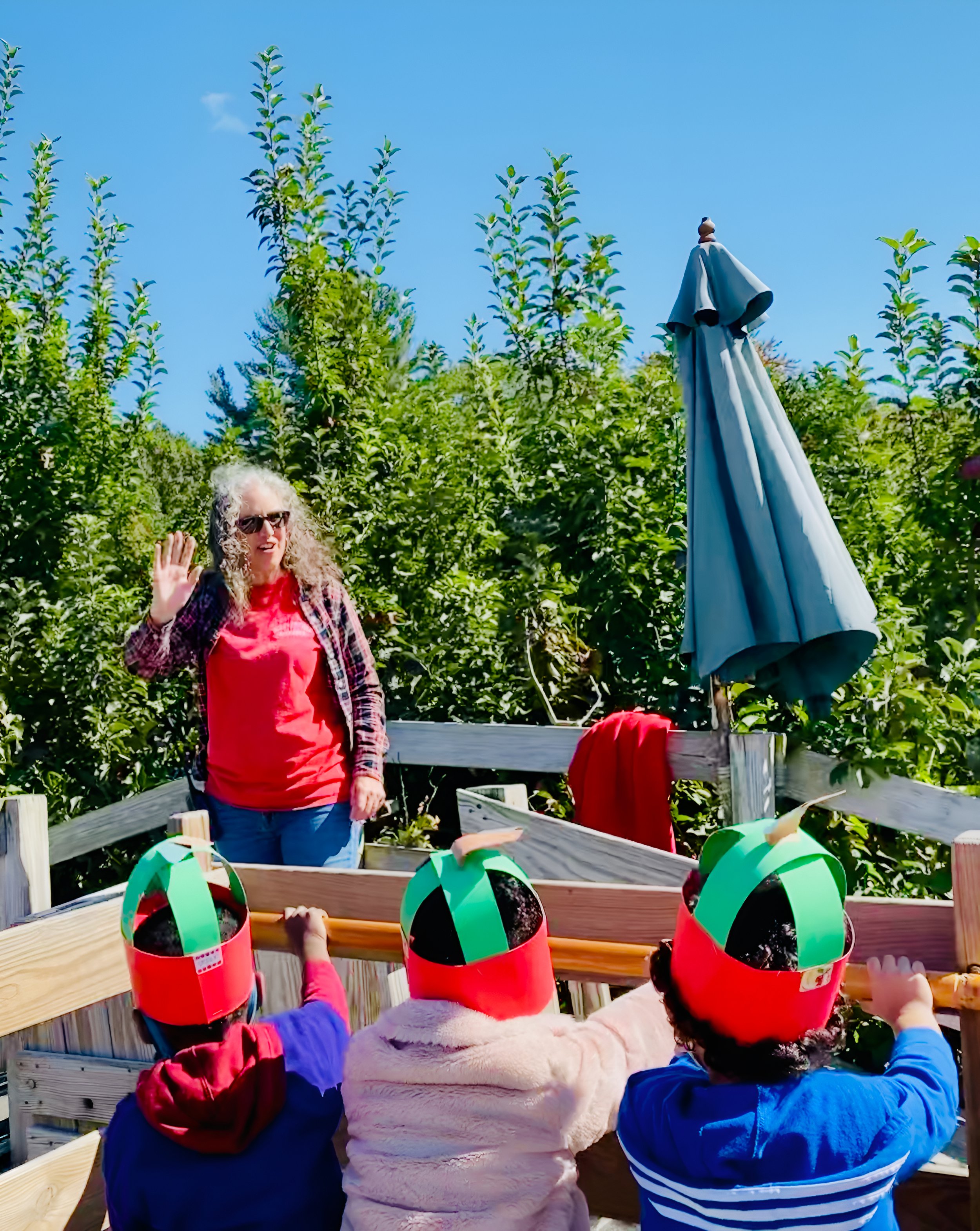 Children wearing red hats with green and black decorations, reaching out towards a woman dressed as a sheep, standing on a wooden platform outdoors among green trees and blue sky.
