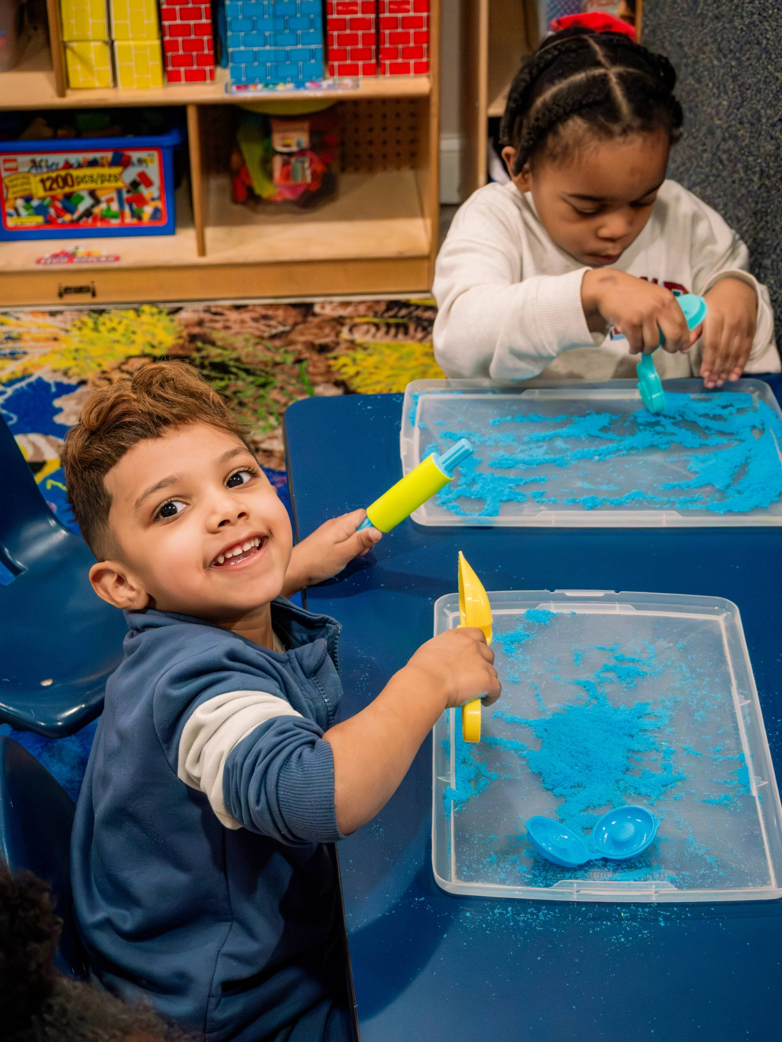 Two young children engaging in a sensory activity with blue colored sand in a classroom setting. One child is smiling at the camera, holding a yellow toy, while the other is focused on using a blue scoop to play with the sand.