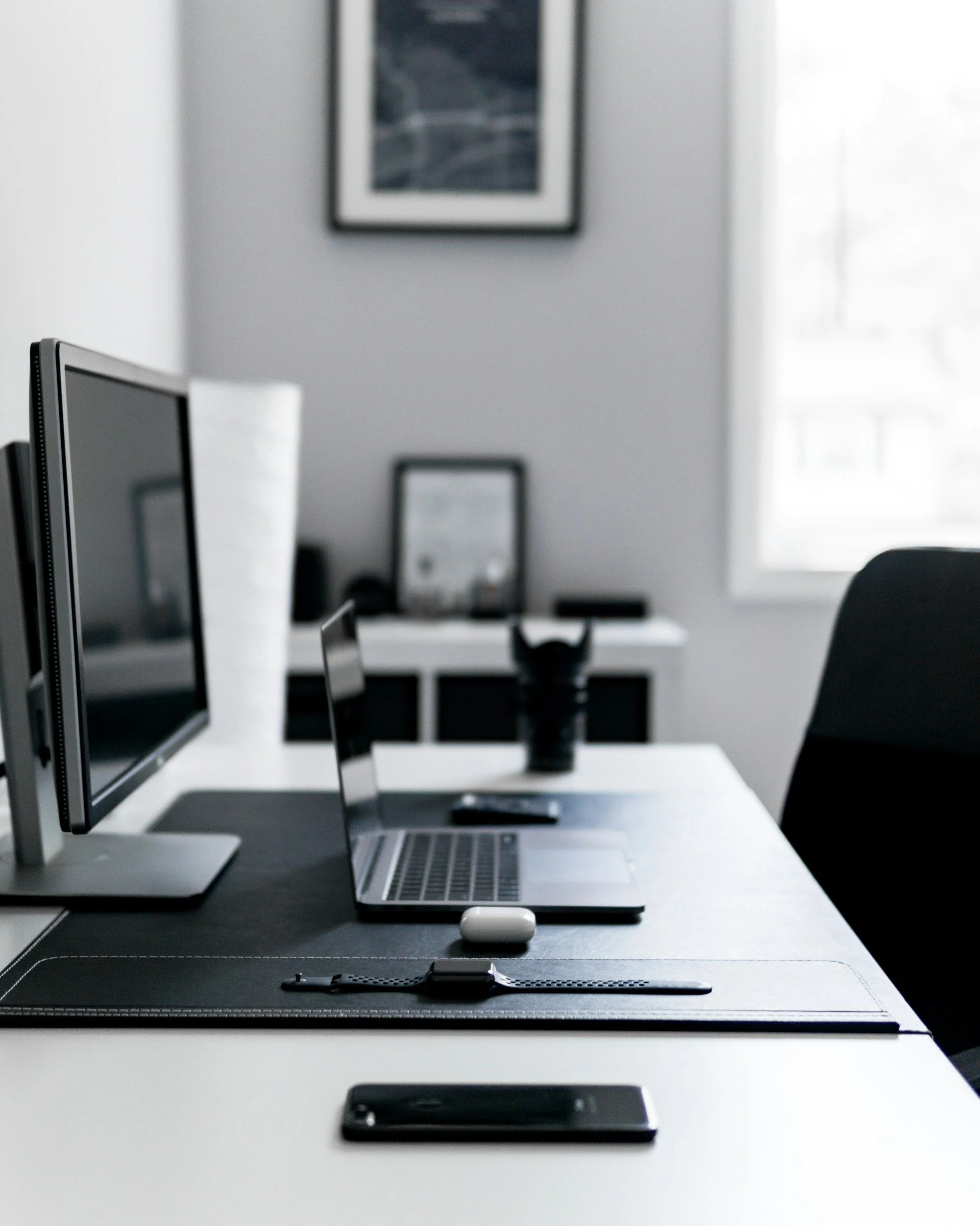 A workspace with a computer monitor, a laptop, a smartphone, and a keyboard on a black desk, with a picture on the wall and a window with white curtains in the background.