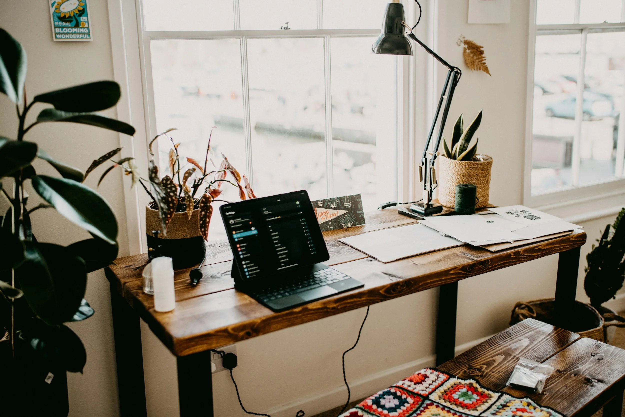 A cozy home office corner with a wooden desk near a window, decorated with several potted plants, a tablet, and papers. Sunlight streams through the window, creating a warm atmosphere.