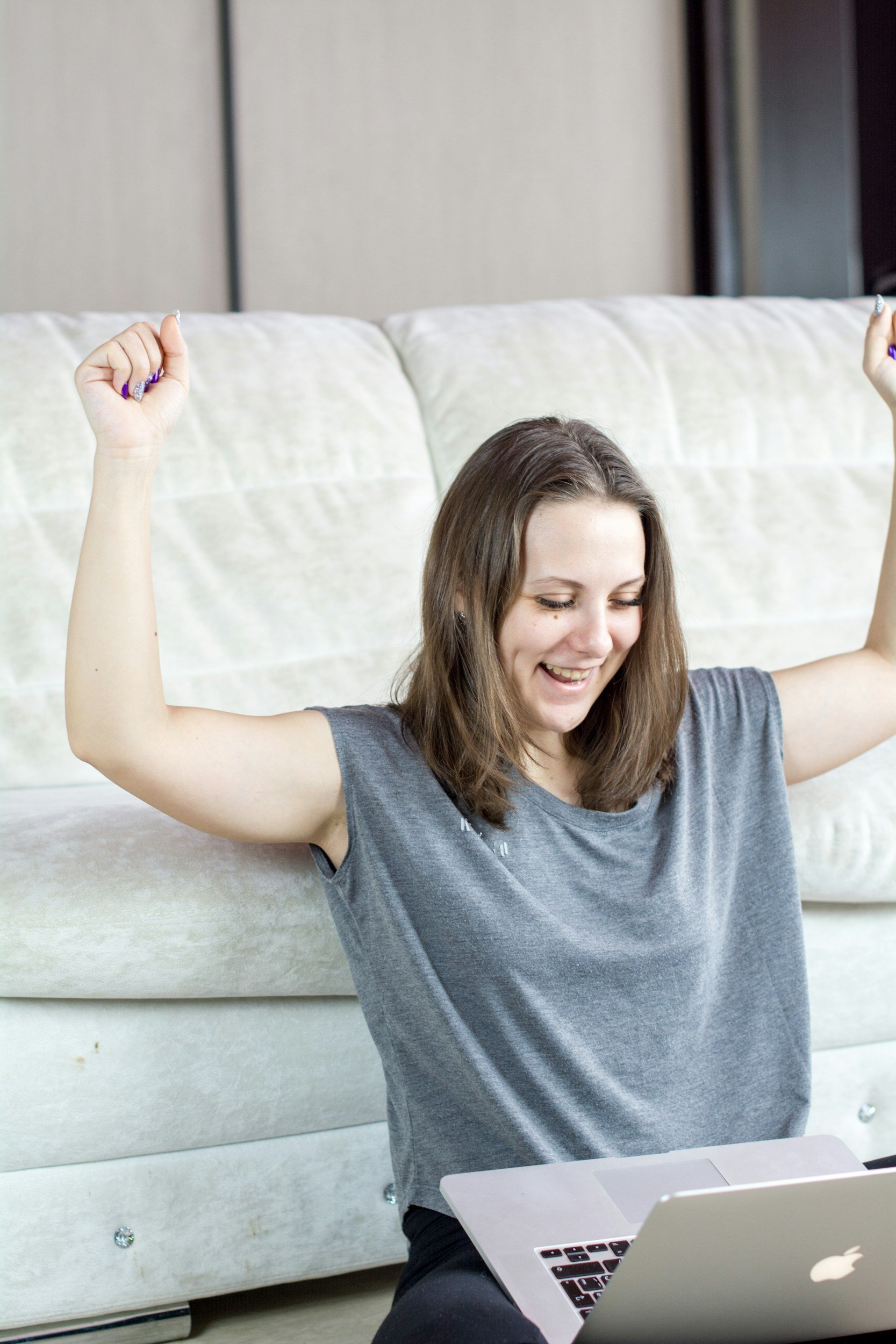 A young woman with brown hair smiling and raising her arms in a celebratory gesture while sitting on a bed with a silver MacBook laptop in front of her.