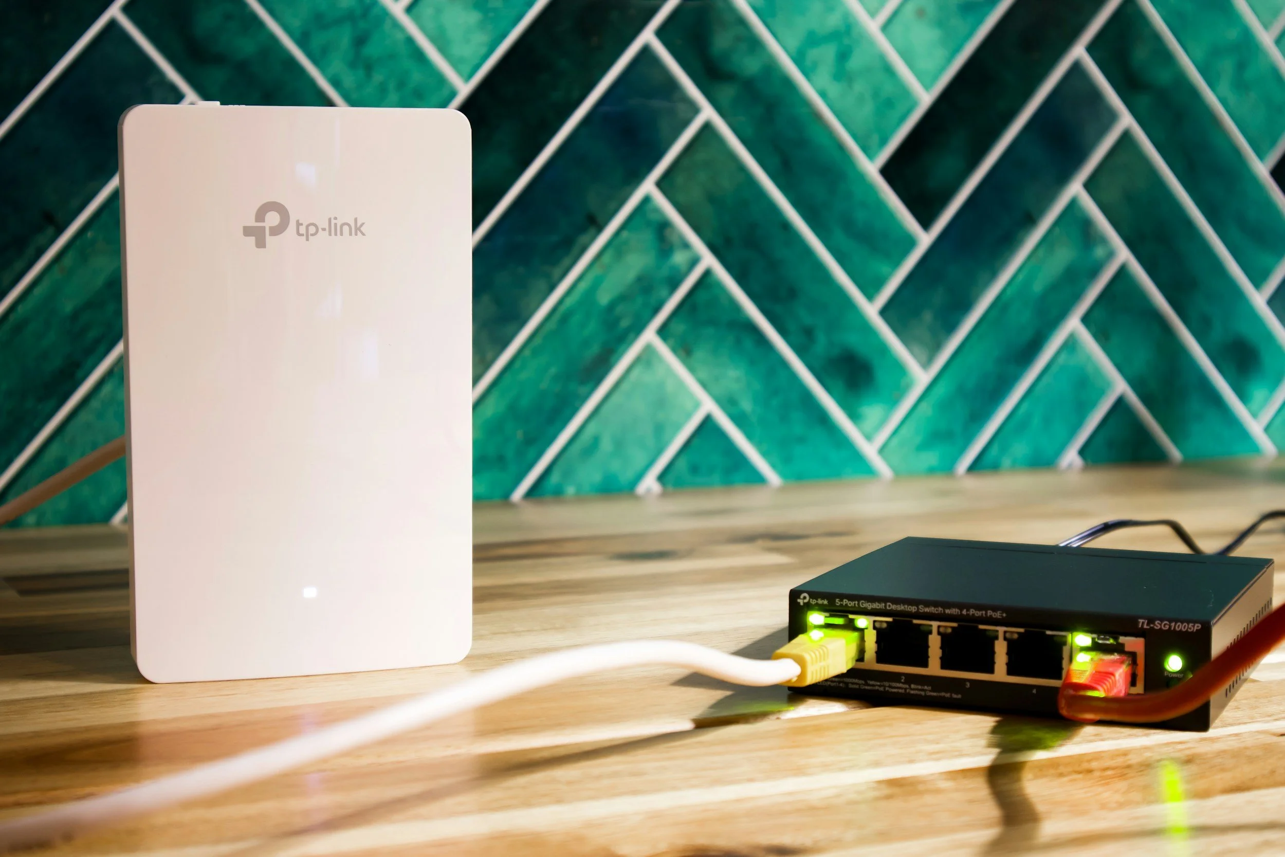 A wireless router with two antennas and a network switch with flashing green lights on a wooden desk. In the background, there is a green and white tiled wall.