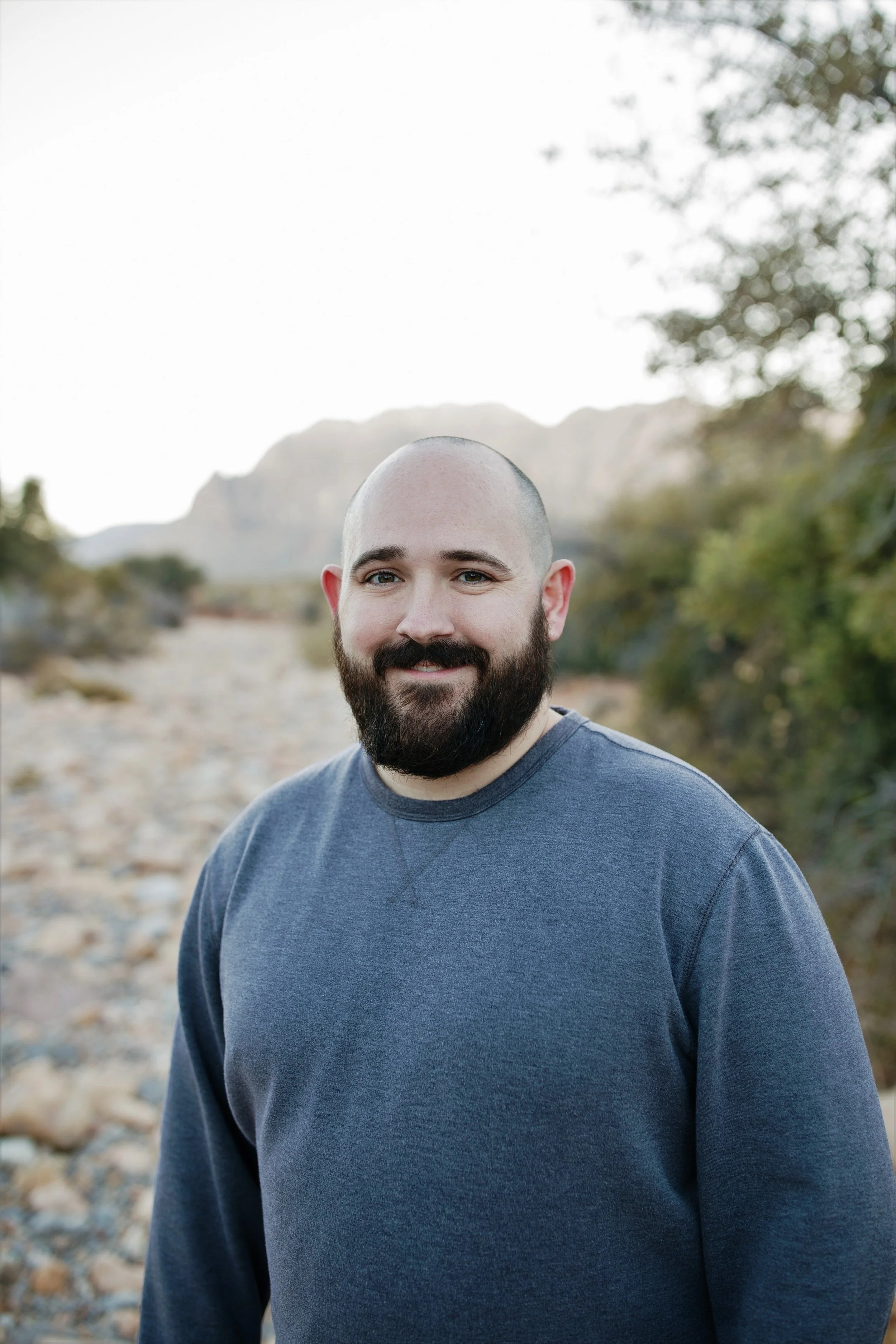 A bearded man with a shaved head wearing a long-sleeve gray shirt, standing outdoors in a rocky natural setting with mountains and trees in the background, smiling at the camera.