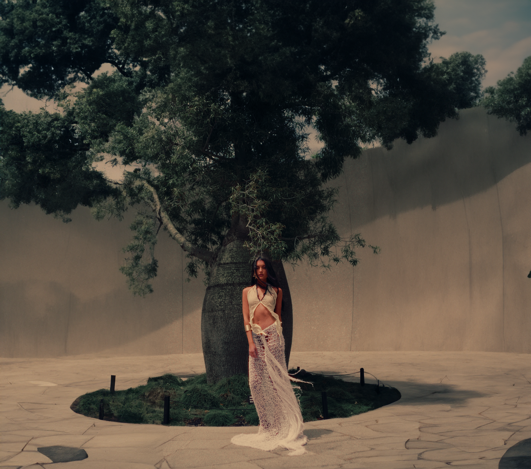 A woman standing in front of a large tree with lush green leaves, wearing a light-colored top and a long, knitted skirt, along with accessories, in an outdoor setting near a concrete wall.