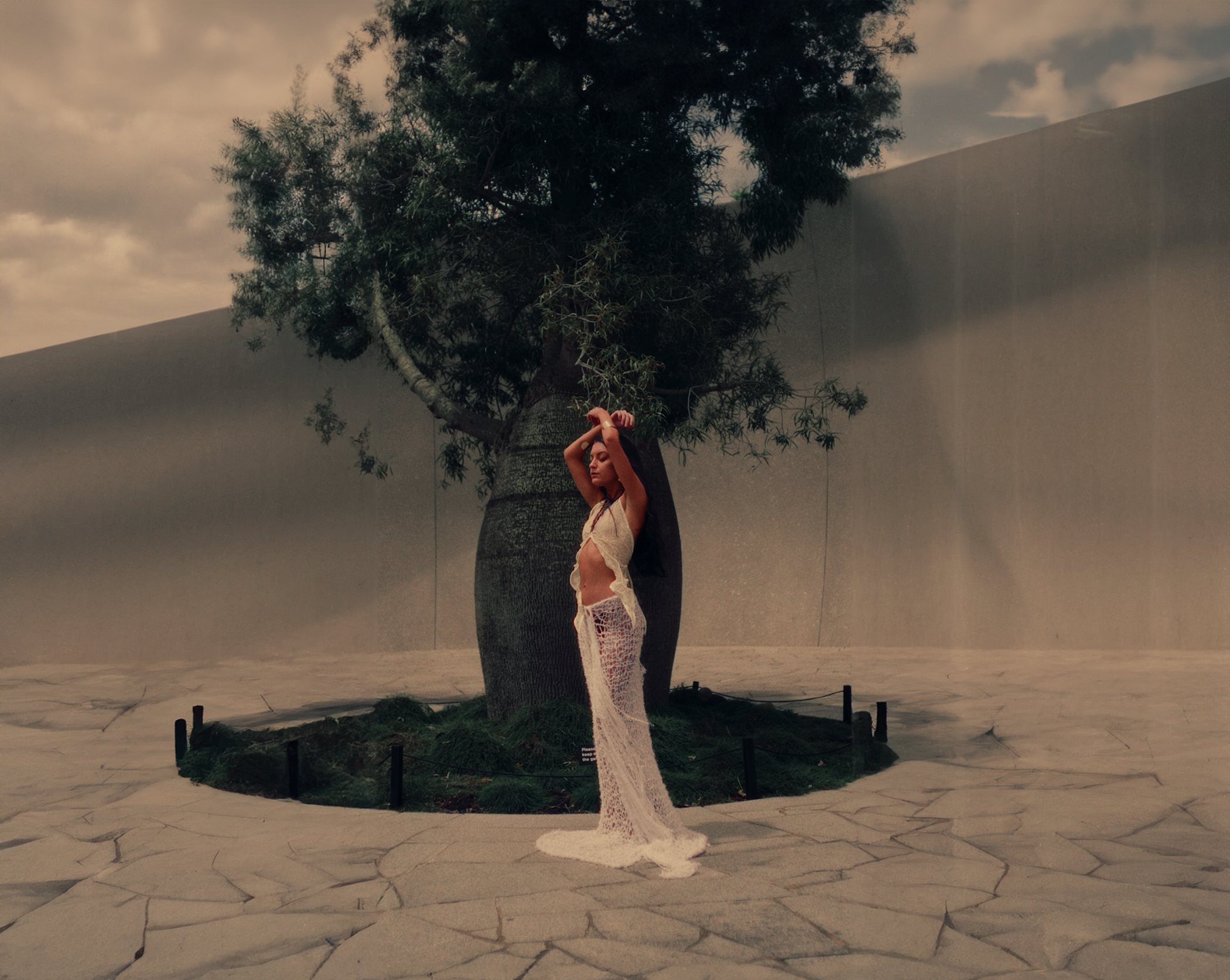 A woman in a long, white, lace dress standing with arms raised in front of a large tree and concrete wall.