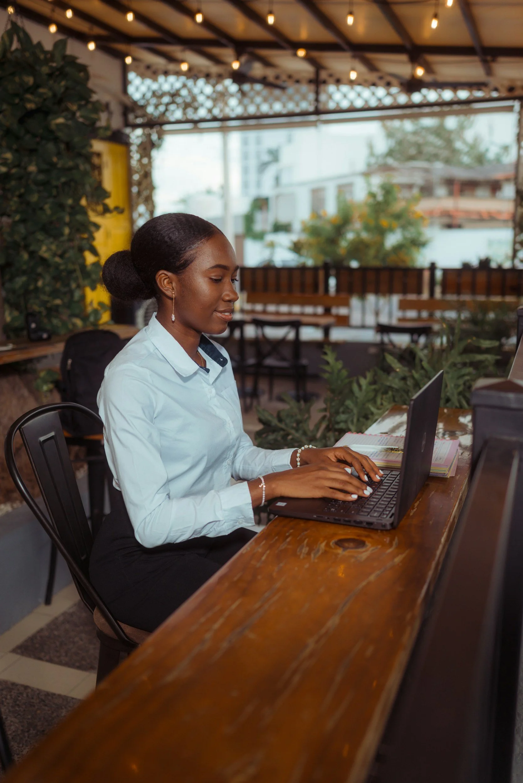 A woman sitting at a wooden table using a laptop in an outdoor or semi-outdoor setting with string lights and plants visible in the background.