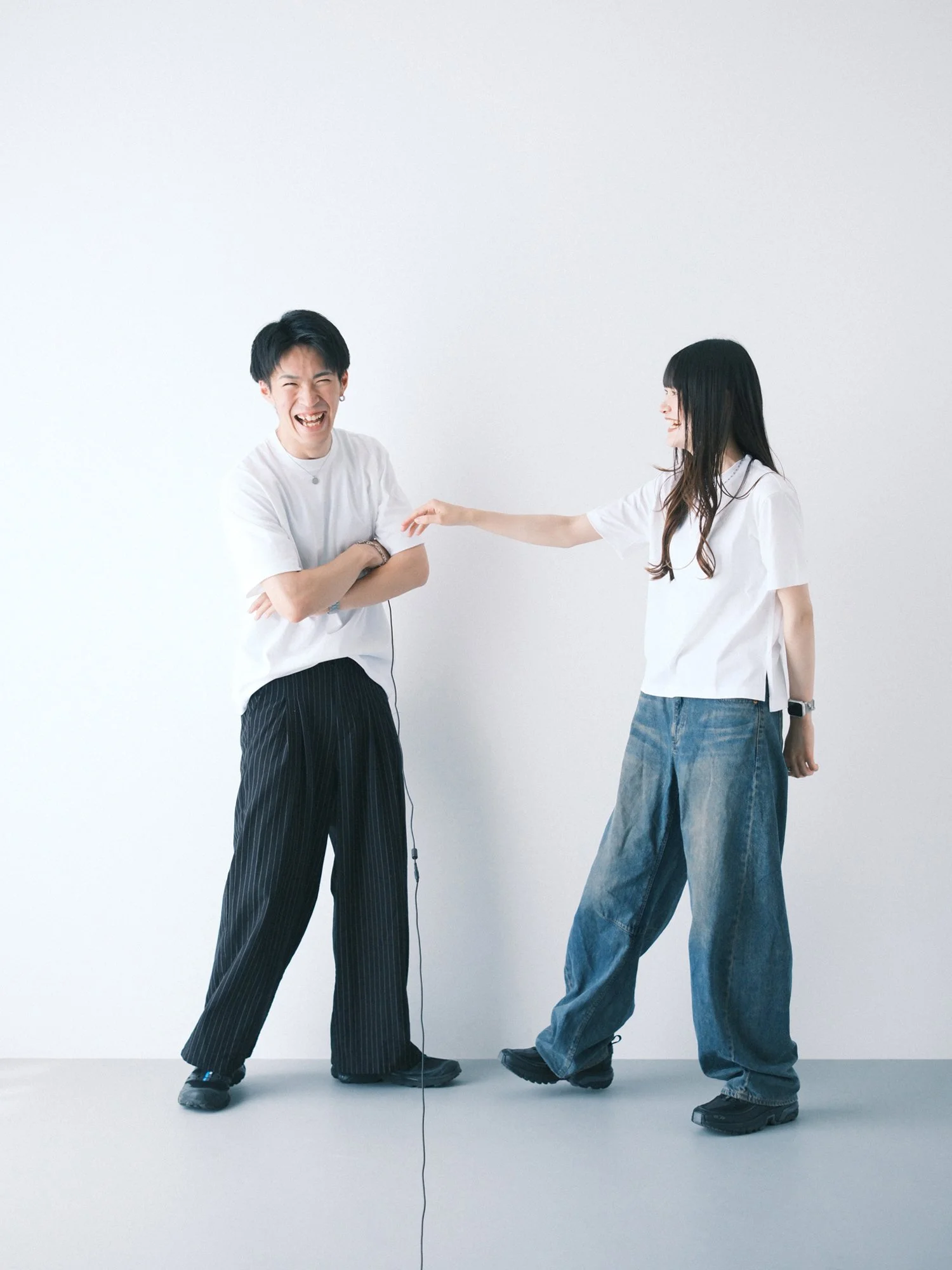 白い背景の前で笑っているカップルが会話している。男性は白いTシャツと縦縞の黒いズボンを着ており、女性は白いシャツとゆったりとした青いジーンズを着ている。男性は腕を組み、女性は伸ばした手で笑顔を見せている。ナチュラルなセルフフォト。