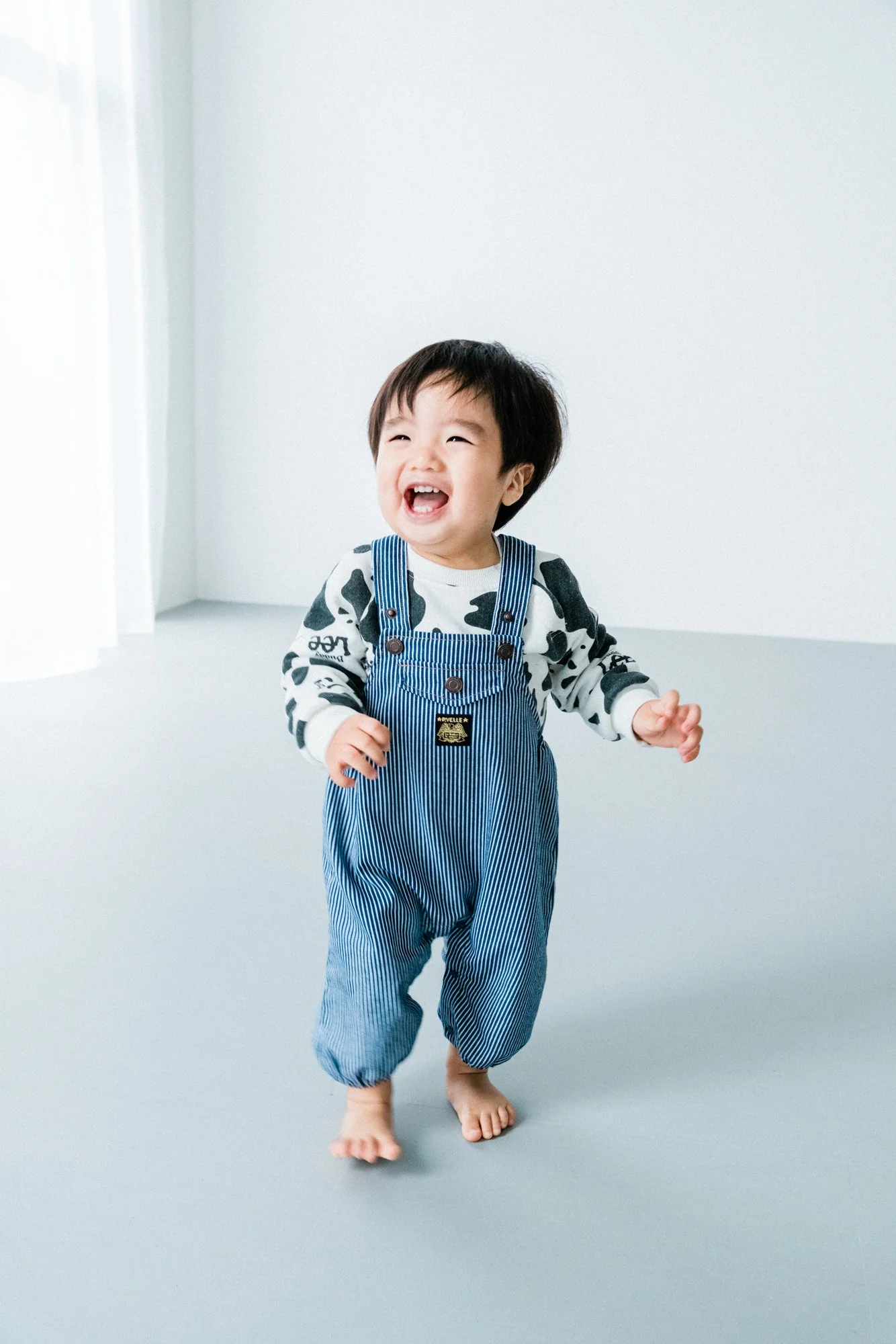 笑って走る小さな子供の男の子。青と白の縞模様のズボンと白と黒の長袖シャツを着ている。裸足で、明るい背景のシンプルなスタジオにいる。