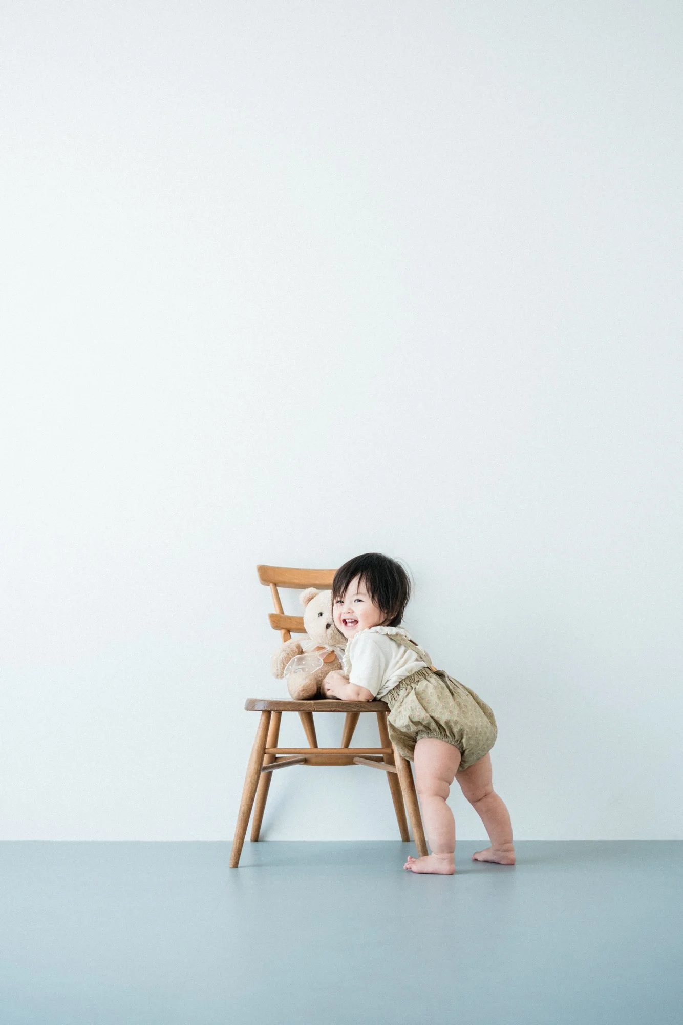 笑顔の子供が椅子の上に座り、ぬいぐるみと一緒に写真撮影をしている様子。背景は白い壁でシンプル。