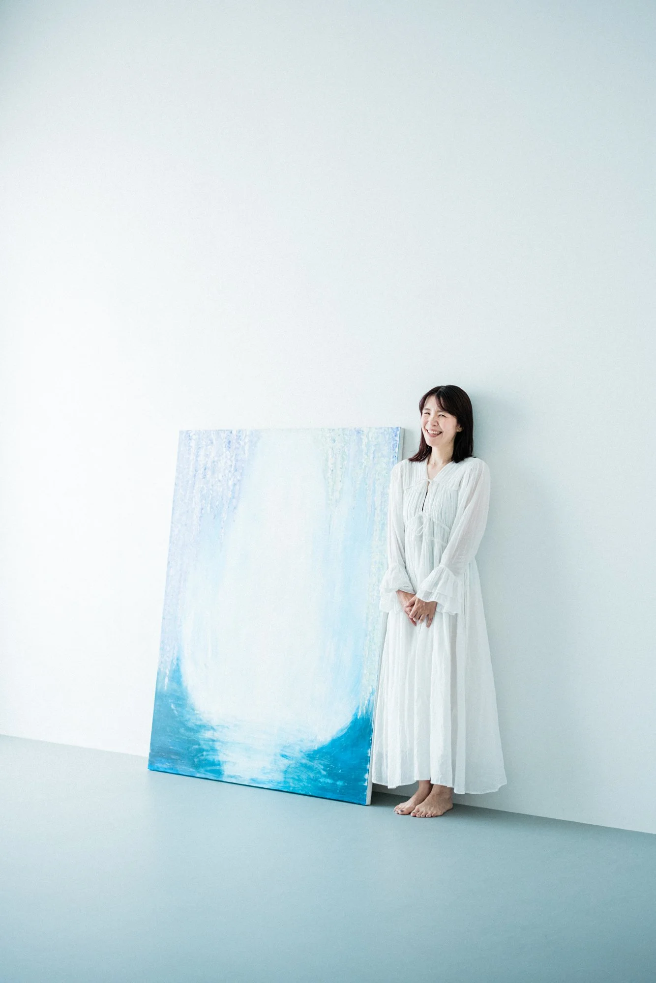 白いワンピースを着た笑顔の女性が無地の壁の前に立ち、床に青と白のアートな抽象画を置いている写真。