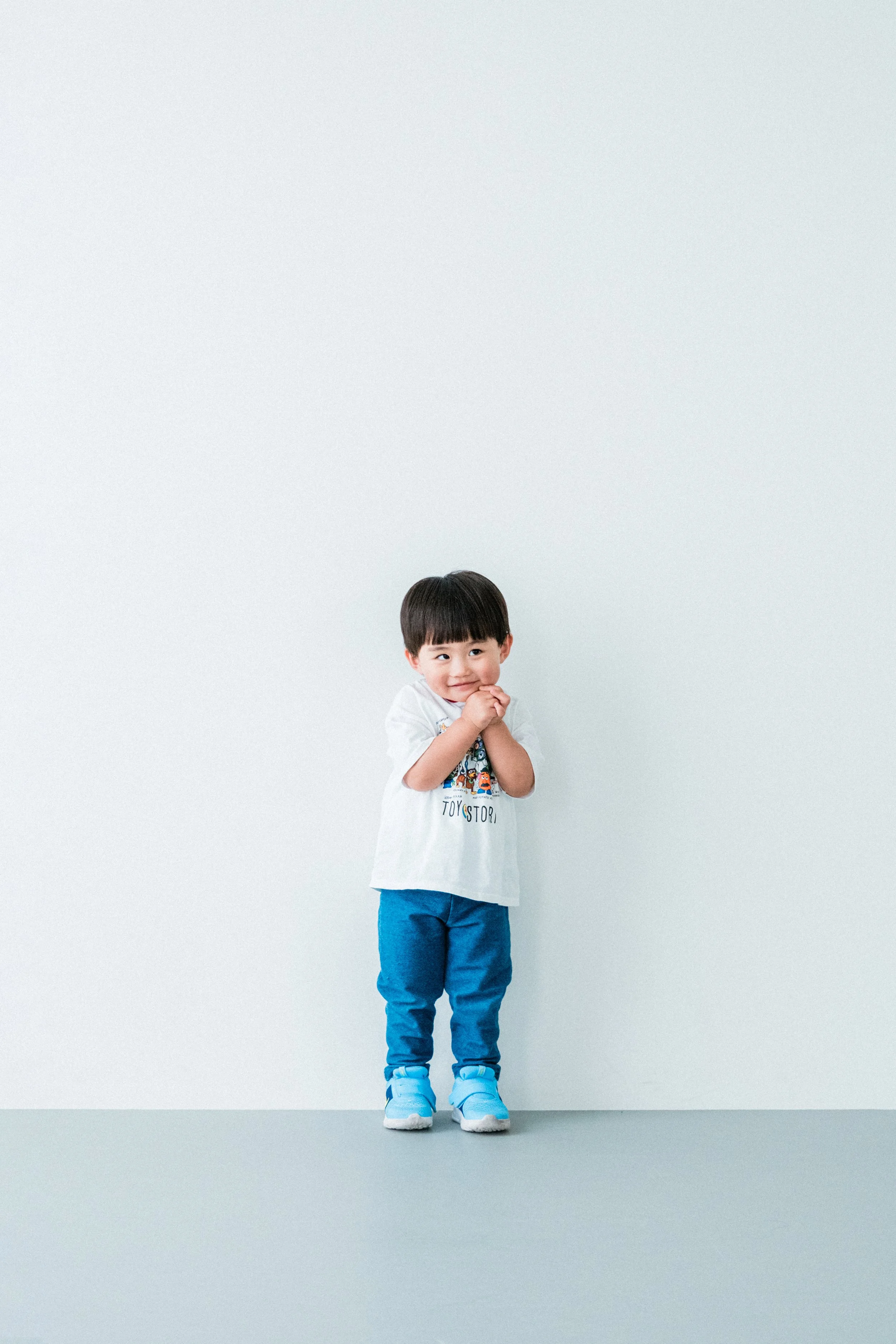 白いTシャツを着て青いパンツとスニーカーを履いた少年が、笑顔で手を口元に当てている。背景はシンプルな白い壁。