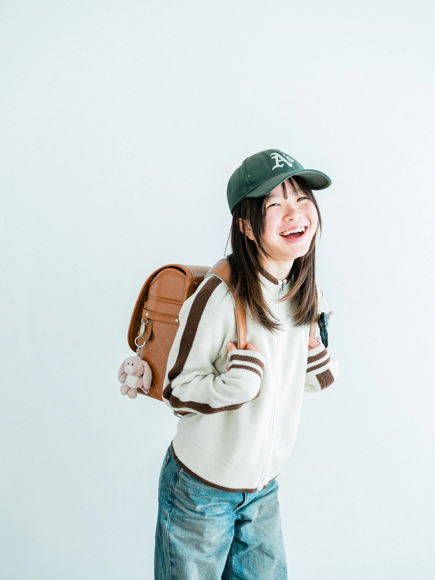 笑顔の女の子がカメラに向かって微笑んでいる。帽子をかぶってランドセルを持ち、カジュアルな服装をしている。入学卒業の記念写真。