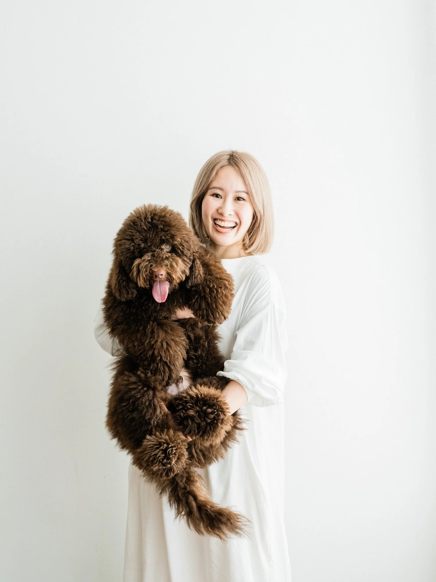白い背景の前に白い服を着た笑顔の女性が、大きな茶色の犬を抱えている写真。大切なペットとの家族写真。