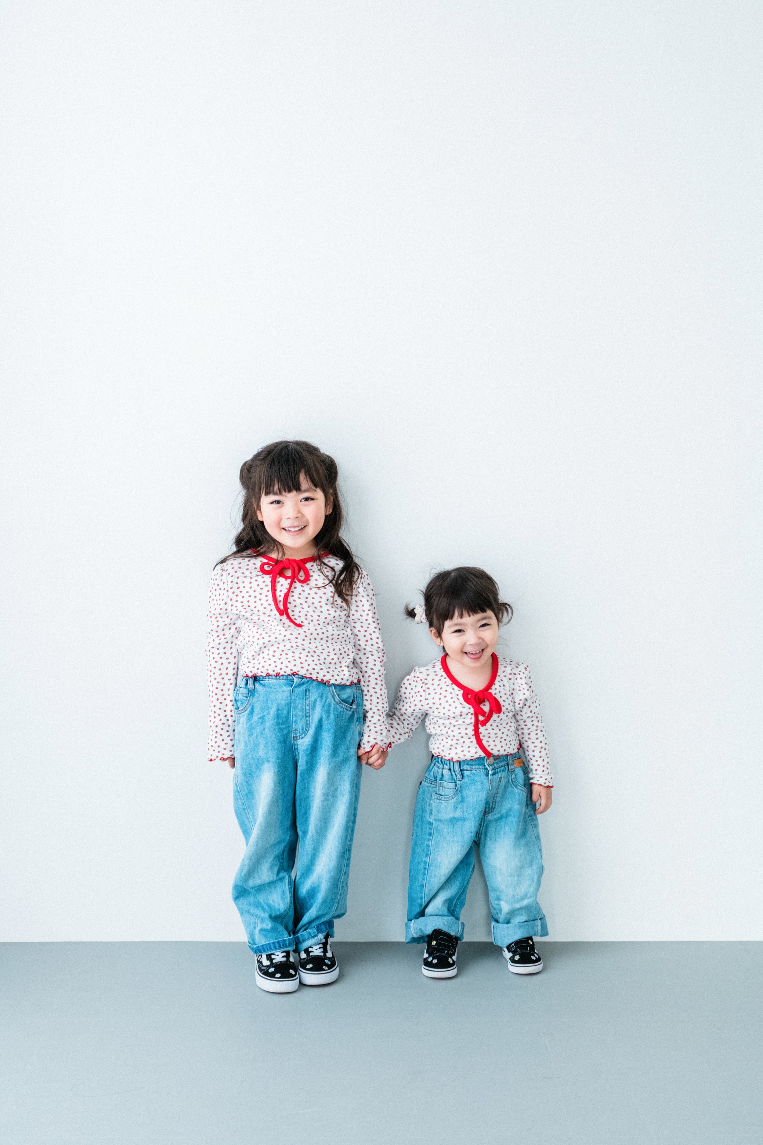 白い背景の前で手をつなぐ二人の姉妹の写真。二人ともお揃いの白地に赤い水玉模様のシャツを着ていて、青いジーンズを履いています。笑顔で並んでいます。