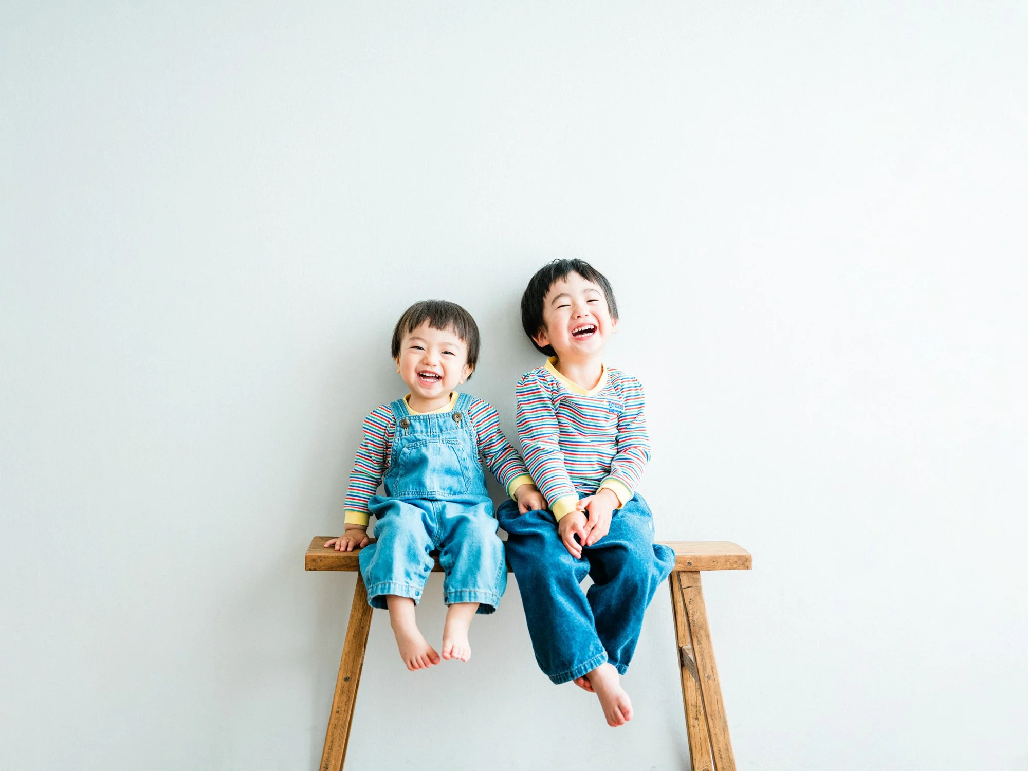 木製のベンチに座る二人の小さな男の子、白い背景の前で笑顔を見せている写真。自然体な兄弟の家族写真。