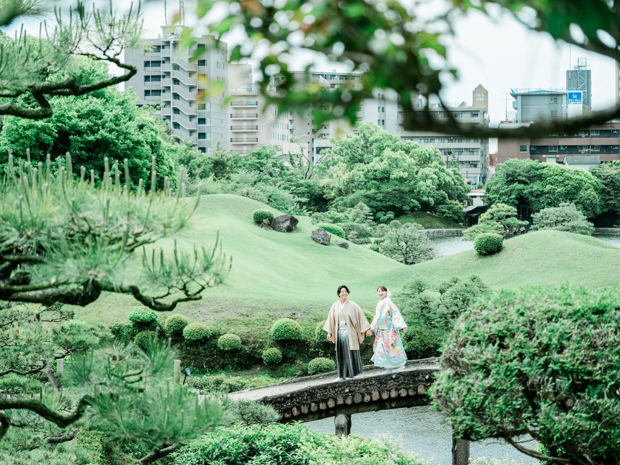 緑の庭園で新郎新婦が和服を着て歩いている光景。背景には都市の建物と木々が見える。自然体なフォトウェディング。