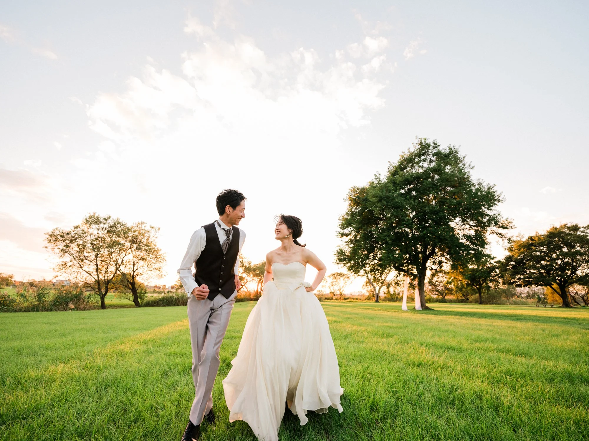 結婚式の新郎新婦が屋外の緑の芝生の公園で笑顔で話している様子。夕日が背景にあり、木々が点在している。出張のフォトウェディング。