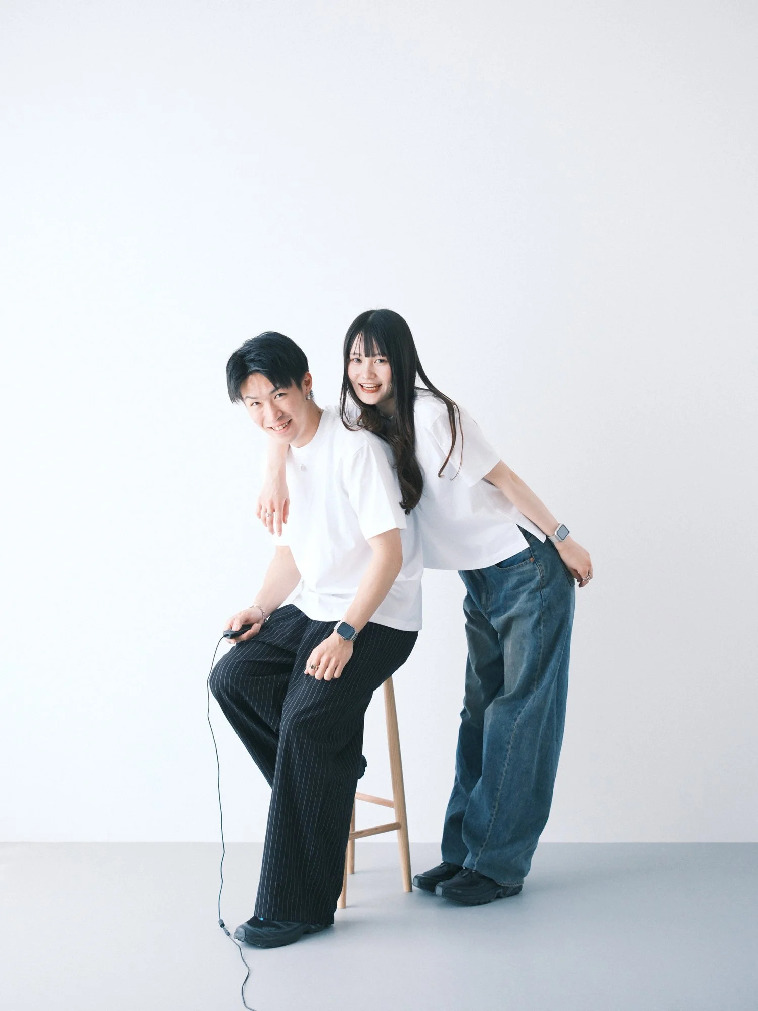 白いTシャツを着た男の人が座っており、黒い縦縞のパンツを穿いている。彼の後ろに白いTシャツを着た女の人が立ち、二人とも笑顔でポーズをとっている。カップルのセルフフォト。