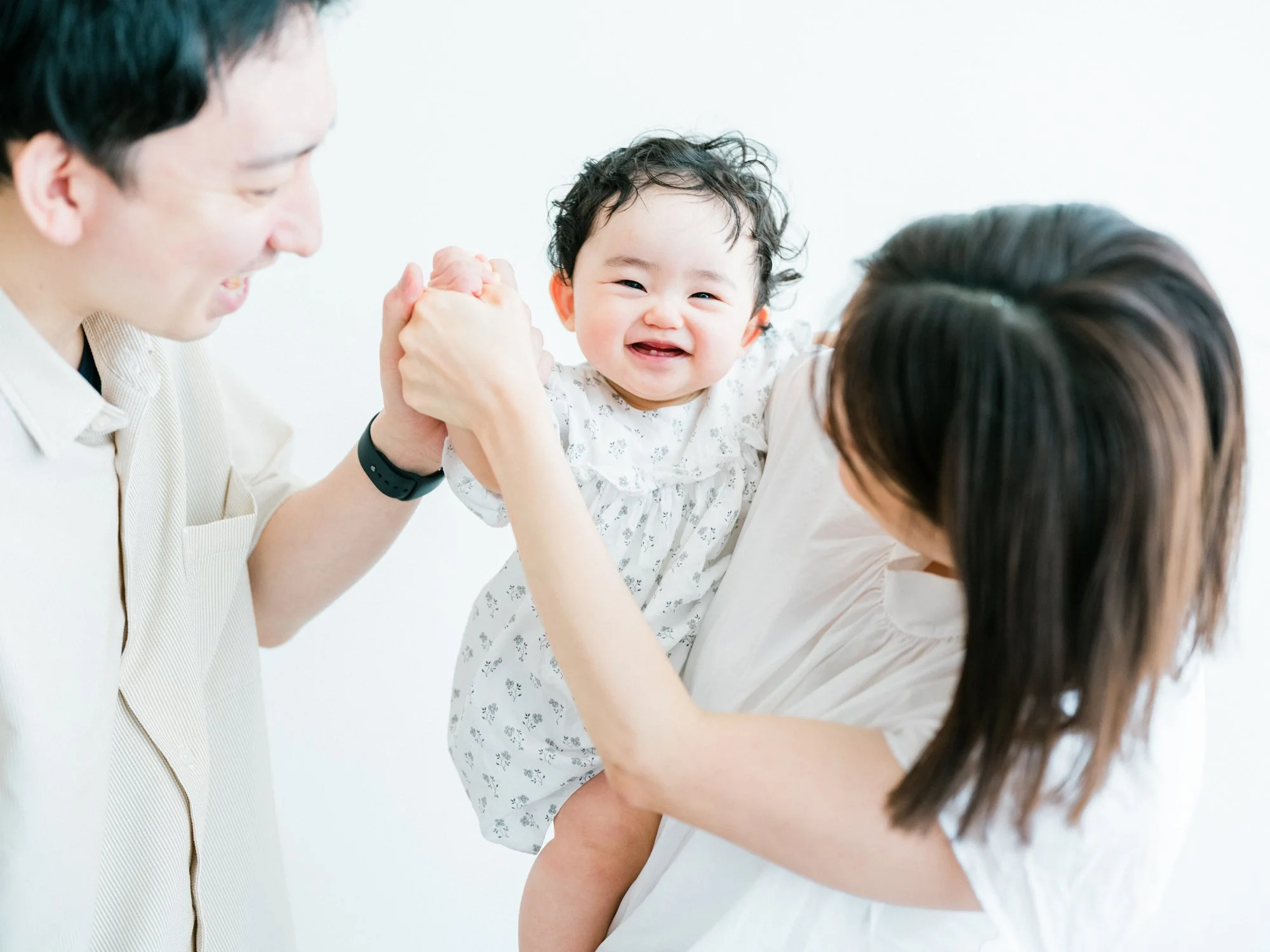 笑顔の赤ちゃんと両親が手を繋いでいる自然体な家族写真。