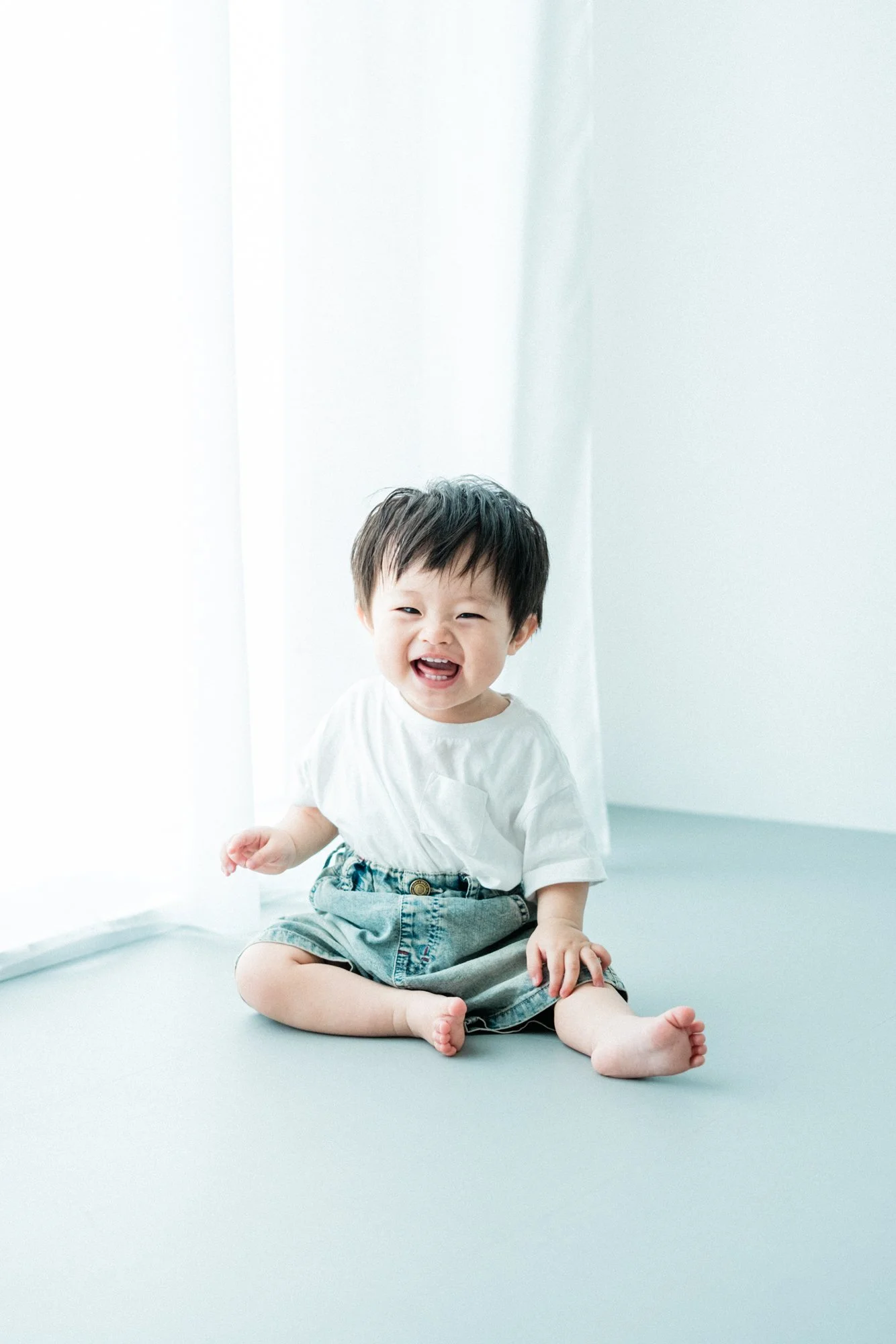 笑顔の幼い1歳の男の子が白いシャツとデニムパンツをはいて座っている。背景は白い壁と窓から自然光が差し込んでいる。