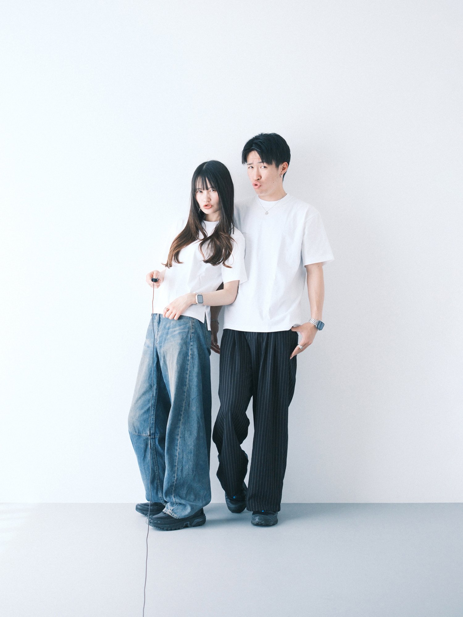 白い背景の前に立つカップル。女性はジーンズをはき、男性はズボンをはいている。二人とも白いTシャツを着ている。カジュアルなセルフフォト。