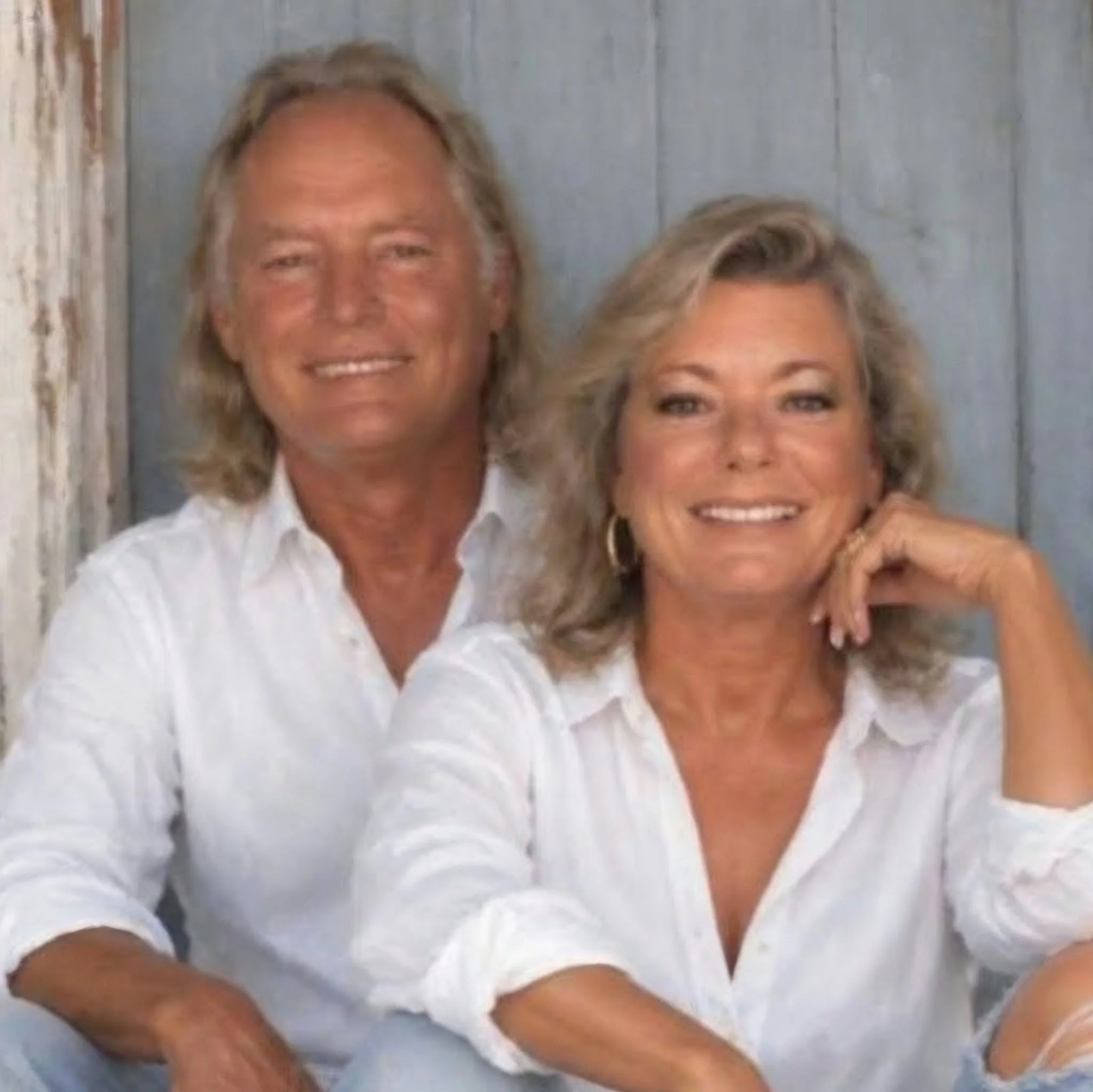 Two people wearing white shirts, smiling, standing close together, with one person's arm reaching into the frame, in a room with a light-colored wooden wall.
