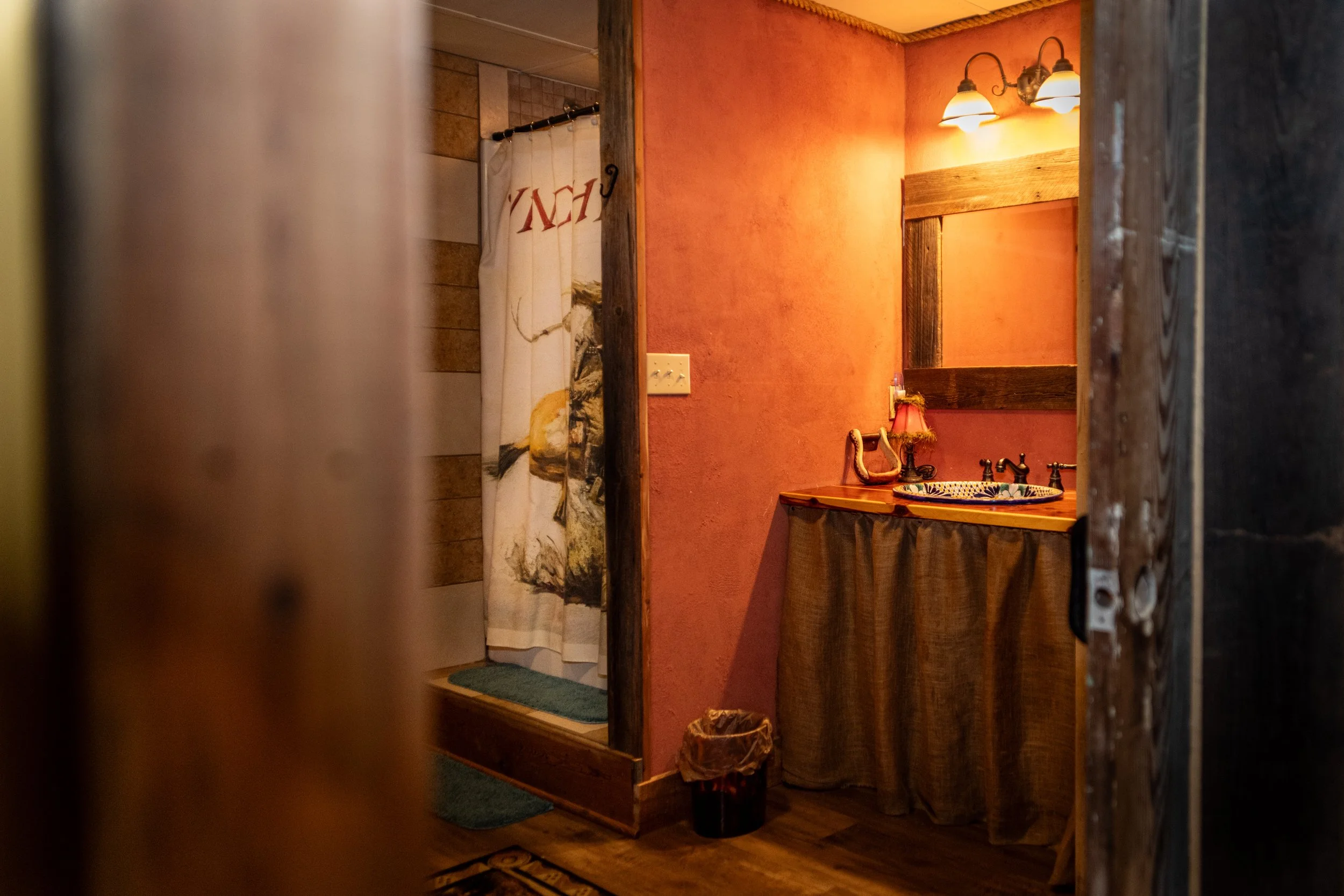 A rustic-style bathroom with warm orange walls, a wooden-framed mirror, and a light fixture with two lamps. There is a sink with a colorful basin, a small lamp with a fringed shade, and a towel hanging beneath the mirror. The photo appears to be take