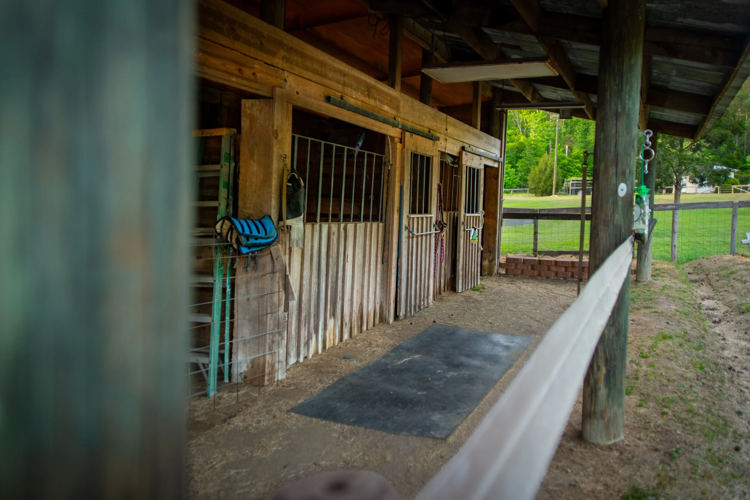 View of a horse stable with wooden doors and a fenced outdoor area, surrounded by green trees and grass.