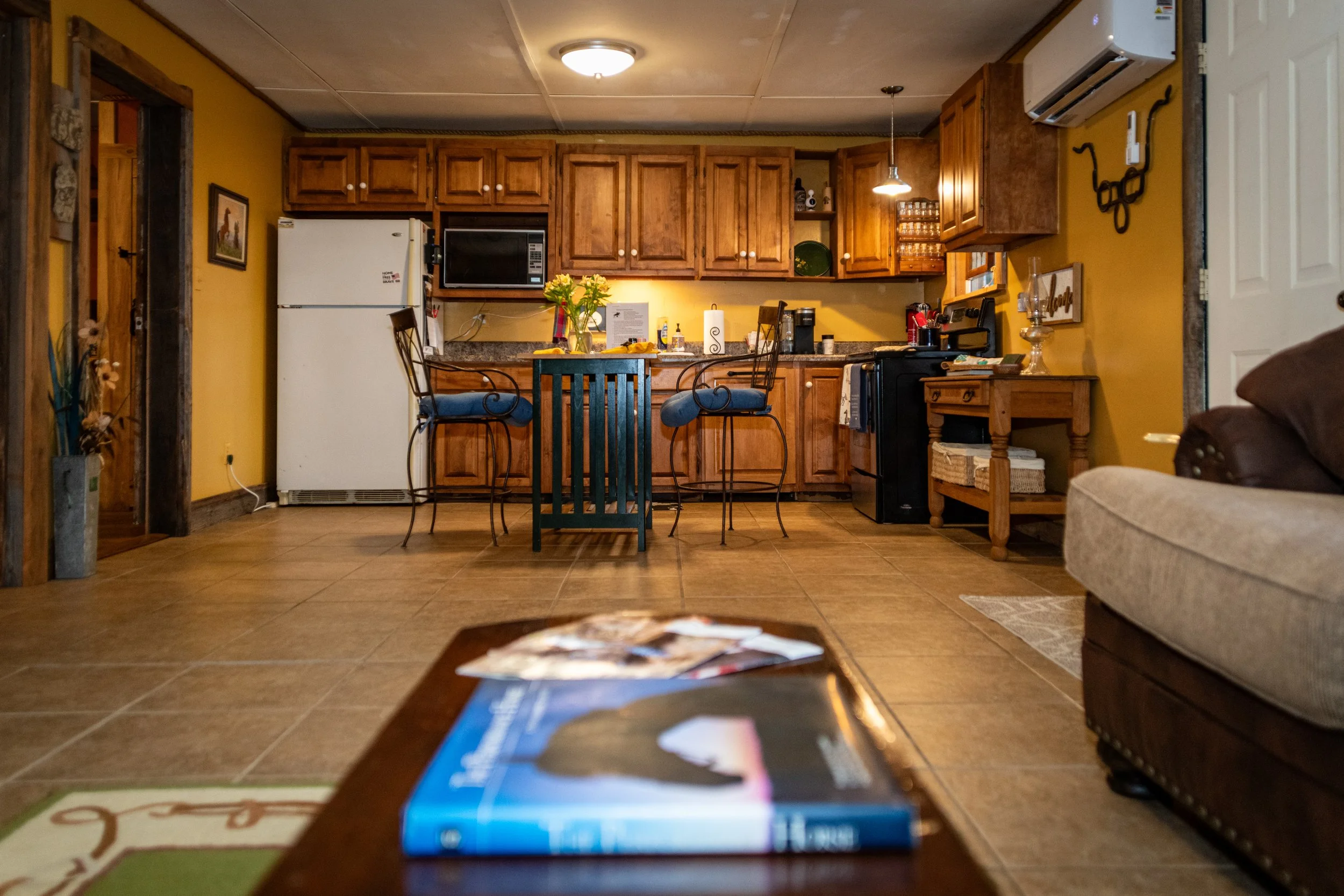 View of a cozy kitchen with wooden cabinets, a white refrigerator, a microwave, and a small dining area with two chairs. The photo is taken from a low angle with a magazine on a coffee table in the foreground.