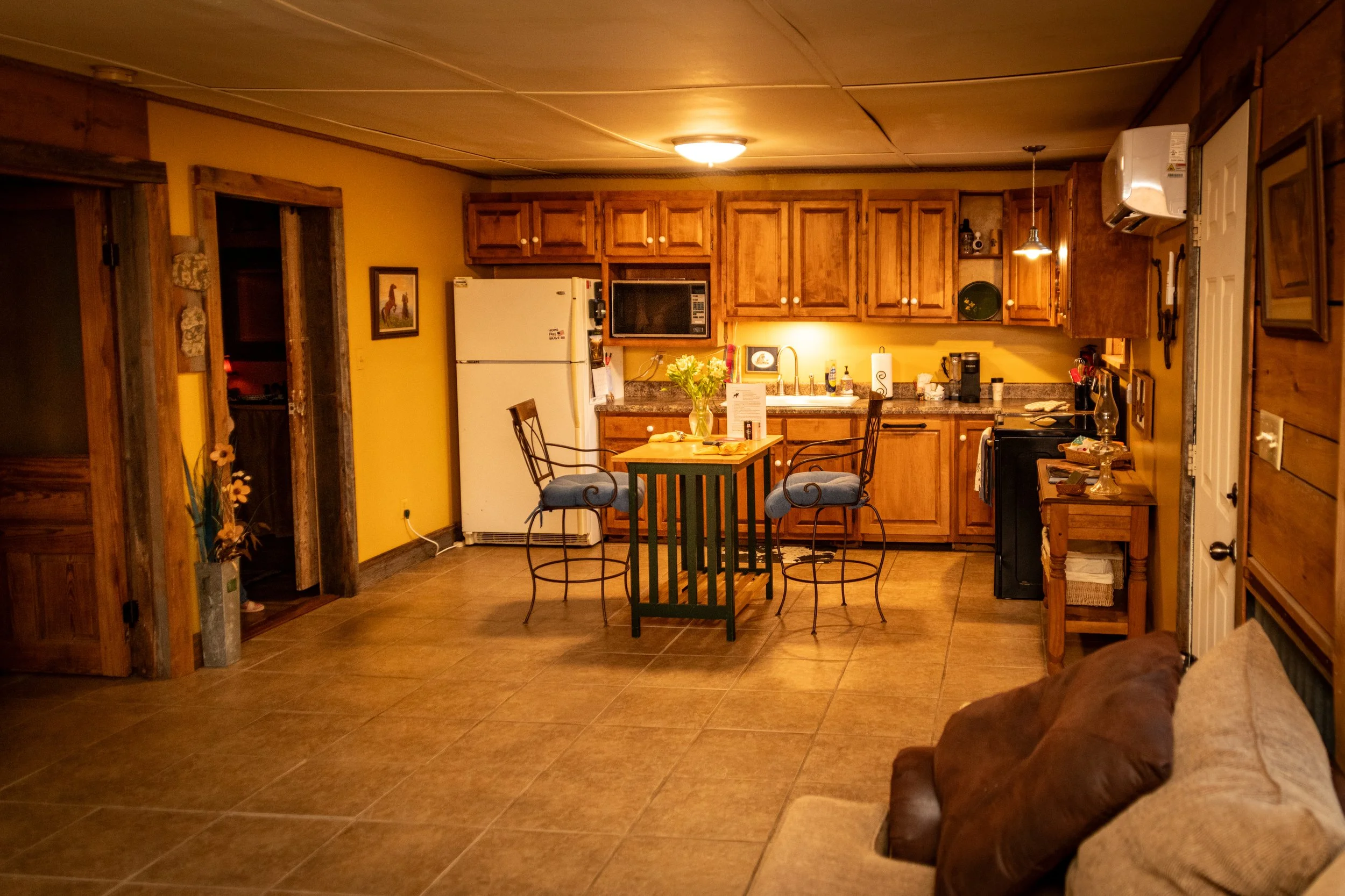 A cozy kitchen with wooden cabinets, a white refrigerator, a microwave, and a small dining table with four chairs. There are decorative items on the walls and counter, and a vase of flowers on the table.