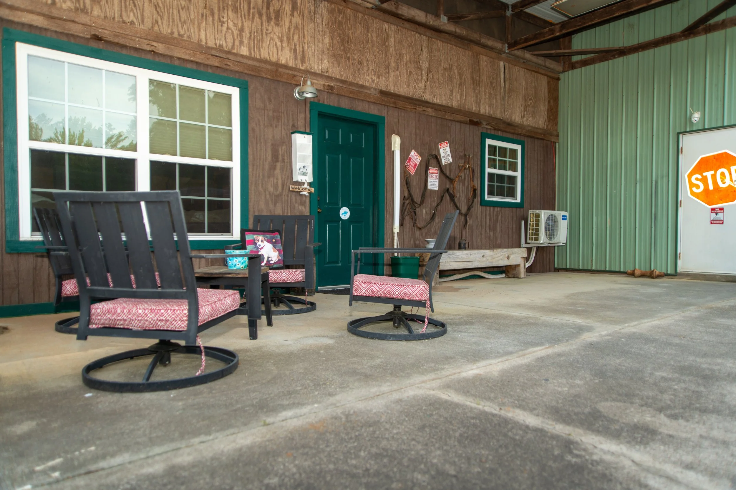 View of outdoor patio area with black metal chairs with red and white cushions, wooden walls, a dark green door, two windows with green trim, outdoor wall lamp, an air conditioning unit, and a stop sign.